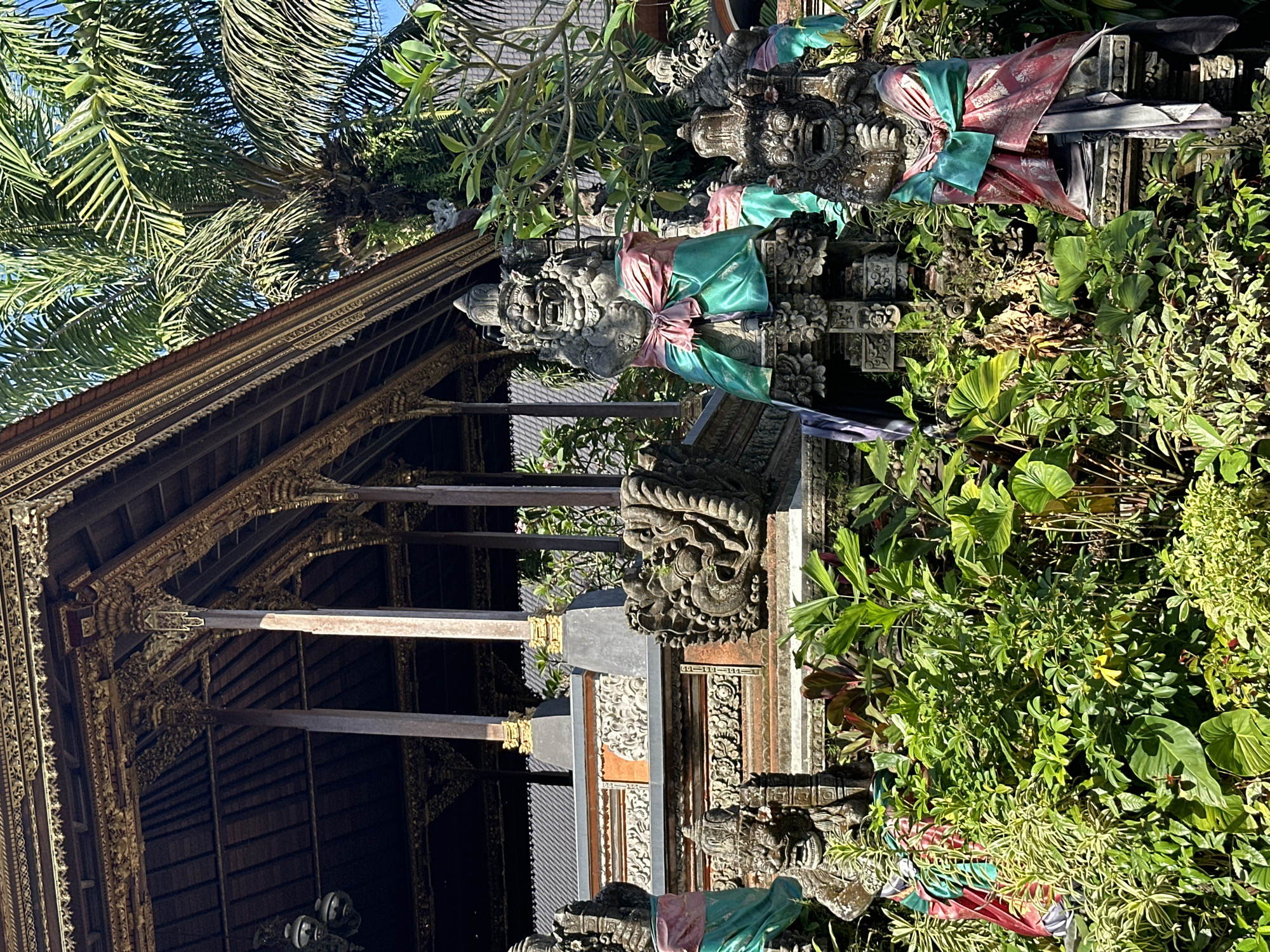 This is an exterior, eye-level, medium shot of Balinese statues and temple architecture in Ubud District, Indonesia. The statue in the foreground is made of grey stone and is draped in a red and green fabric. To the left of this statue is another similar statue, also draped in red and green fabric. In the background, there is a traditional Balinese temple structure with intricate golden carvings and a thatched roof. Lush green vegetation surrounds the statues and temple. The sky is clear and blue. The lighting suggests it is daytime, with bright sunlight casting shadows. There are no people visible in the image.