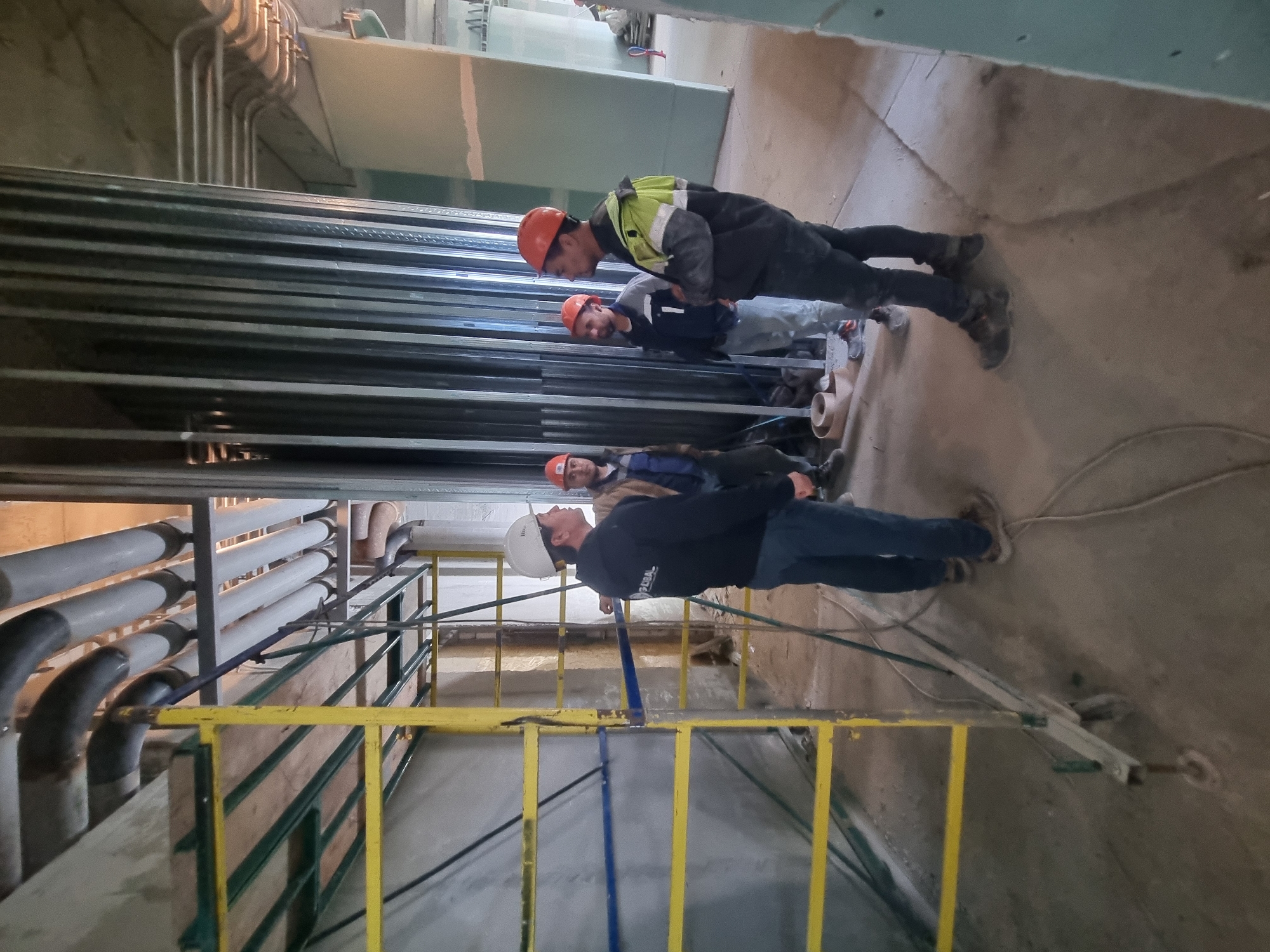 A group of four men, wearing hard hats, stand in what appears to be an industrial or construction site, potentially in Sochi, Russia. They stand near stacked metal framing materials, possibly wall studs or similar construction components. Behind the men are various pipes and a yellow and green metal scaffolding-like structure. Some drywall sheets are visible further in the background. The floor is concrete. One man wears a white hard hat, while the other three wear orange hard hats. One man wears a high visibility vest. The men appear to be observing something in the scene.