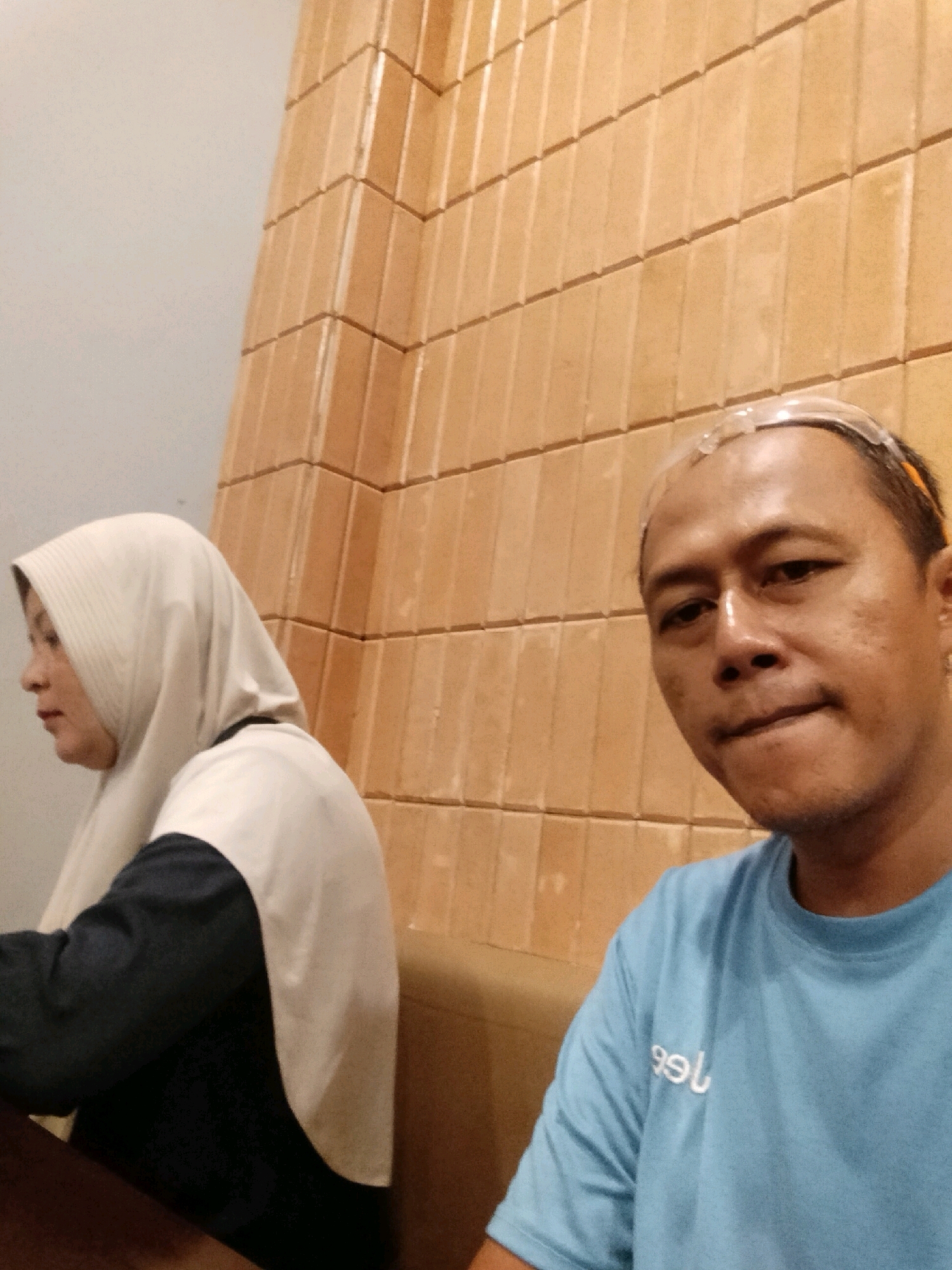 In Arang Limbung, Indonesia, a selfie is taken of two individuals against a textured, orange-brown wall. On the right, a man with a light blue t-shirt and clear protective eyewear on his forehead looks directly at the camera. His t-shirt has white lettering. To the left, a woman wearing a light-colored hijab and a dark shirt with a light gray overlay is partially visible, looking to her left. The background consists of stacked, rectangular tiles with visible grout lines. The lighting is dim, suggesting an indoor setting.