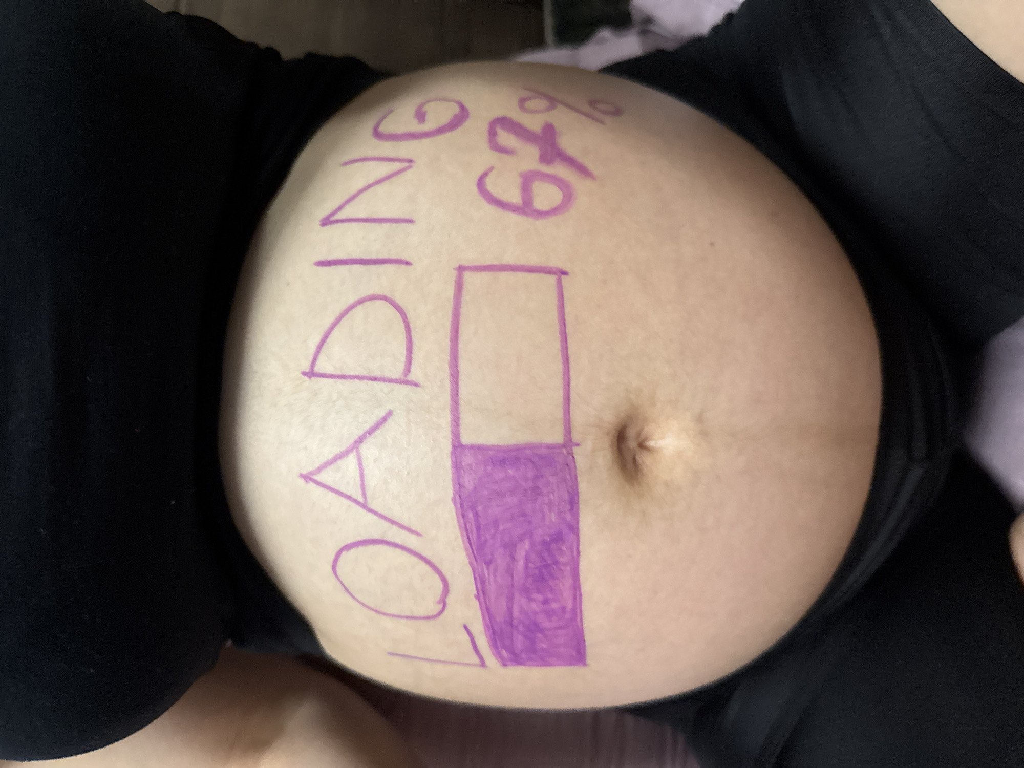 The image shows a close-up of a pregnant person's belly, decorated with purple marker. The belly is round and prominently displays the word "LOADING" written vertically. To the right of the word, "67%" is written, with a purple bar graph indicating progress. The person is wearing black clothing, which covers their torso and legs. The background is blurred and appears to be indoors, with hints of purple fabric. The overall tone of the image is playful and expectant, likely capturing a moment during the pregnancy. There are no specific cues about the time of day, weather, or explicit location within Tbilisi, Georgia, beyond what is inferred from the subject matter and the provided location context.