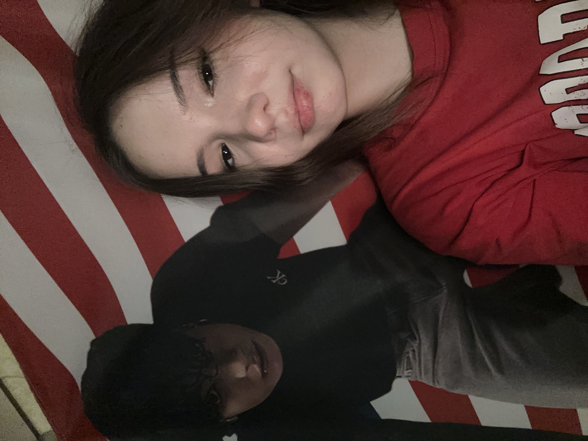 In Naberezhnye Chelny, Russia, a young woman with brown hair and fair skin, wearing a red t-shirt, is taking a selfie. She is positioned in the foreground, looking towards the camera with a neutral expression. Behind her, to the left, is a large poster or print of a young man with dark hair. He is wearing a black t-shirt with a "CK" logo and gray pants. The background behind both individuals consists of red and white stripes, resembling the design of a flag. The lighting appears to be artificial, casting a slight shadow on the woman's face. The overall scene is casual and appears to be indoors.