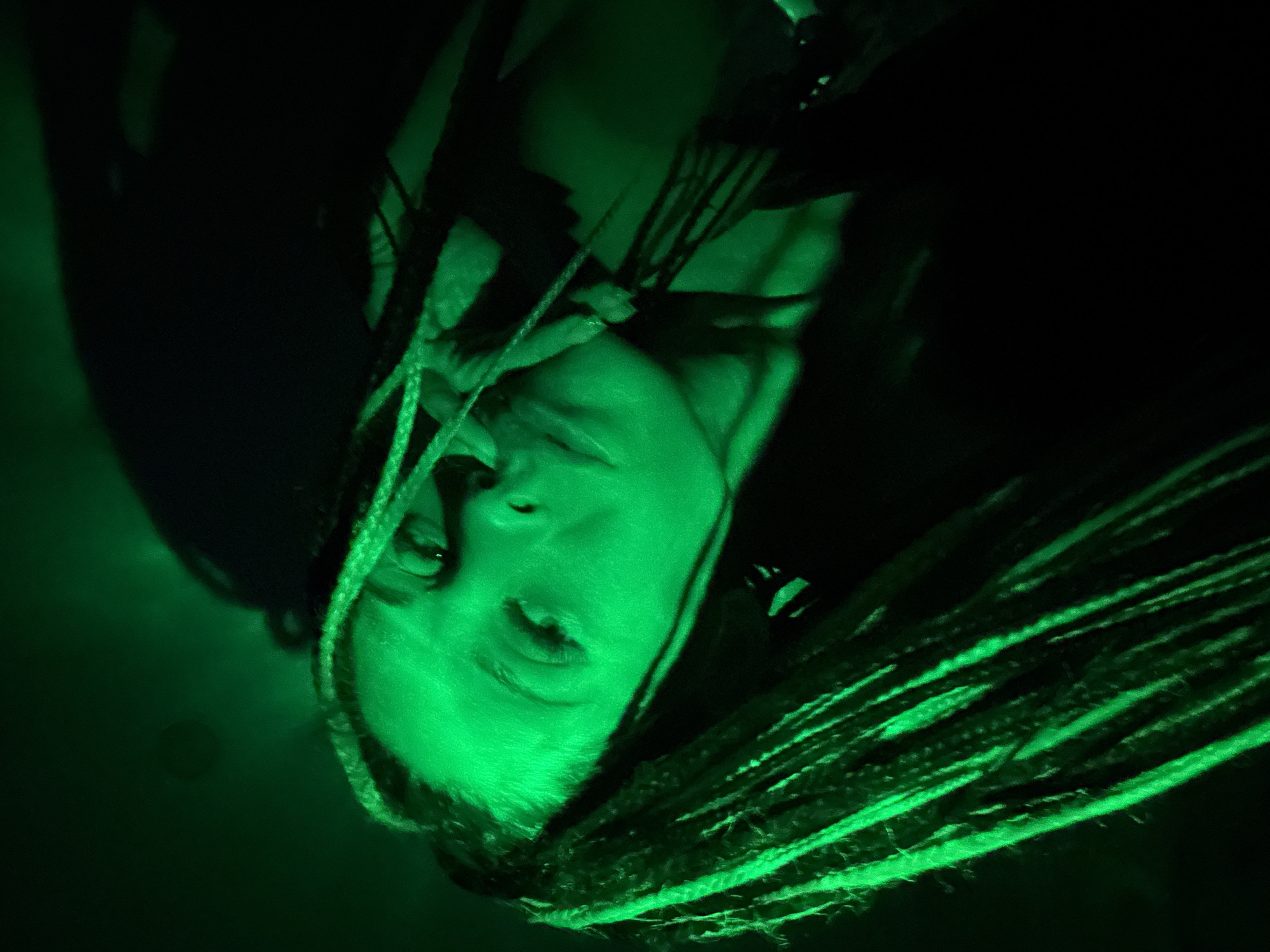A close-up portrait, rotated approximately 90 degrees counter-clockwise, features a person, likely female, under intense green lighting against a dark, indistinct background. The subject's face is positioned on the left side of the frame, tilted upwards, with her eyes open and gazing slightly past the camera. Her lips are subtly parted. Her features are distinctly highlighted by the strong green light, which casts deep shadows, emphasizing the contours of her face. A prominent detail is her long, intricate dreadlocks, some framing her face and others flowing towards the bottom right of the image, also catching the vibrant green illumination. The background is almost entirely black, suggesting a very dark environment or a focused light source. The overall mood is atmospheric and striking, likely taken at night or in a dimly lit indoor setting using artificial green lights. There are no discernible objects beyond the person and her hair, nor any visible text.