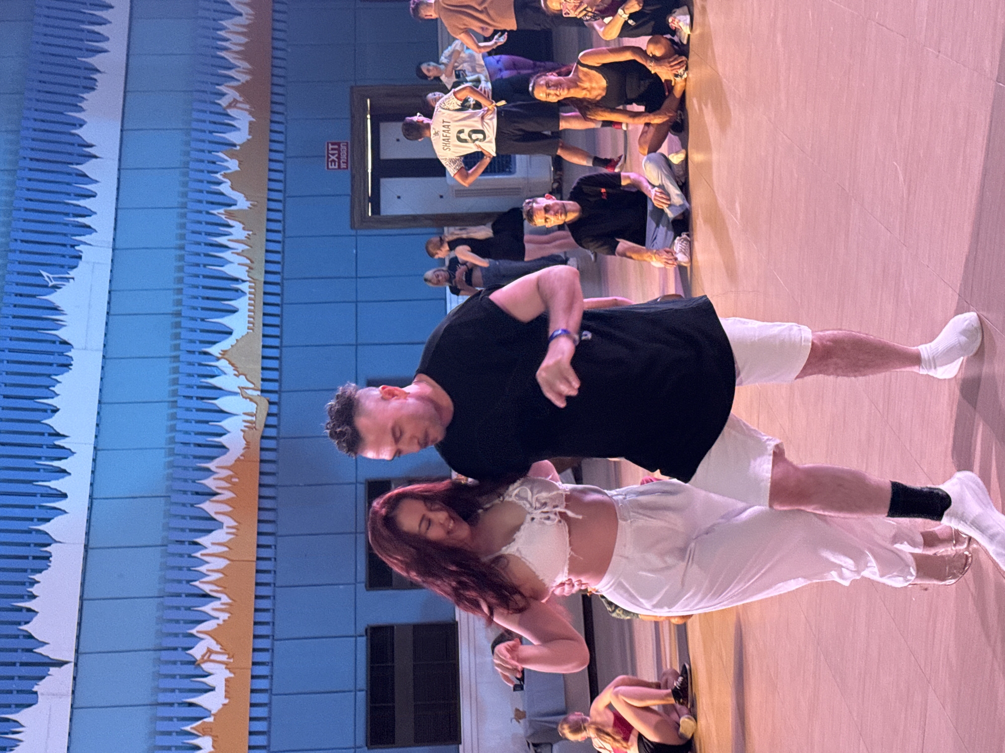 The image captures a group of people in a large room in Pattaya City, Thailand. The scene depicts what appears to be a dance class or rehearsal. A couple stands in the foreground; the woman, with long red hair, wears a white outfit, while the man, with short brown hair, wears a black shirt and white shorts. They seem to be engaged in a dance move, with the woman smiling.
In the background, other people are also seen, some standing, others sitting on the floor. Some are in athletic wear. A man wearing a sports jersey with the name 'SHAFAAT' and the number 6 is visible. An 'EXIT' sign is on the blue wall to the left. The lighting in the room gives off a pink hue, especially on the floor. The building also has blue and beige decorative elements on the facade.