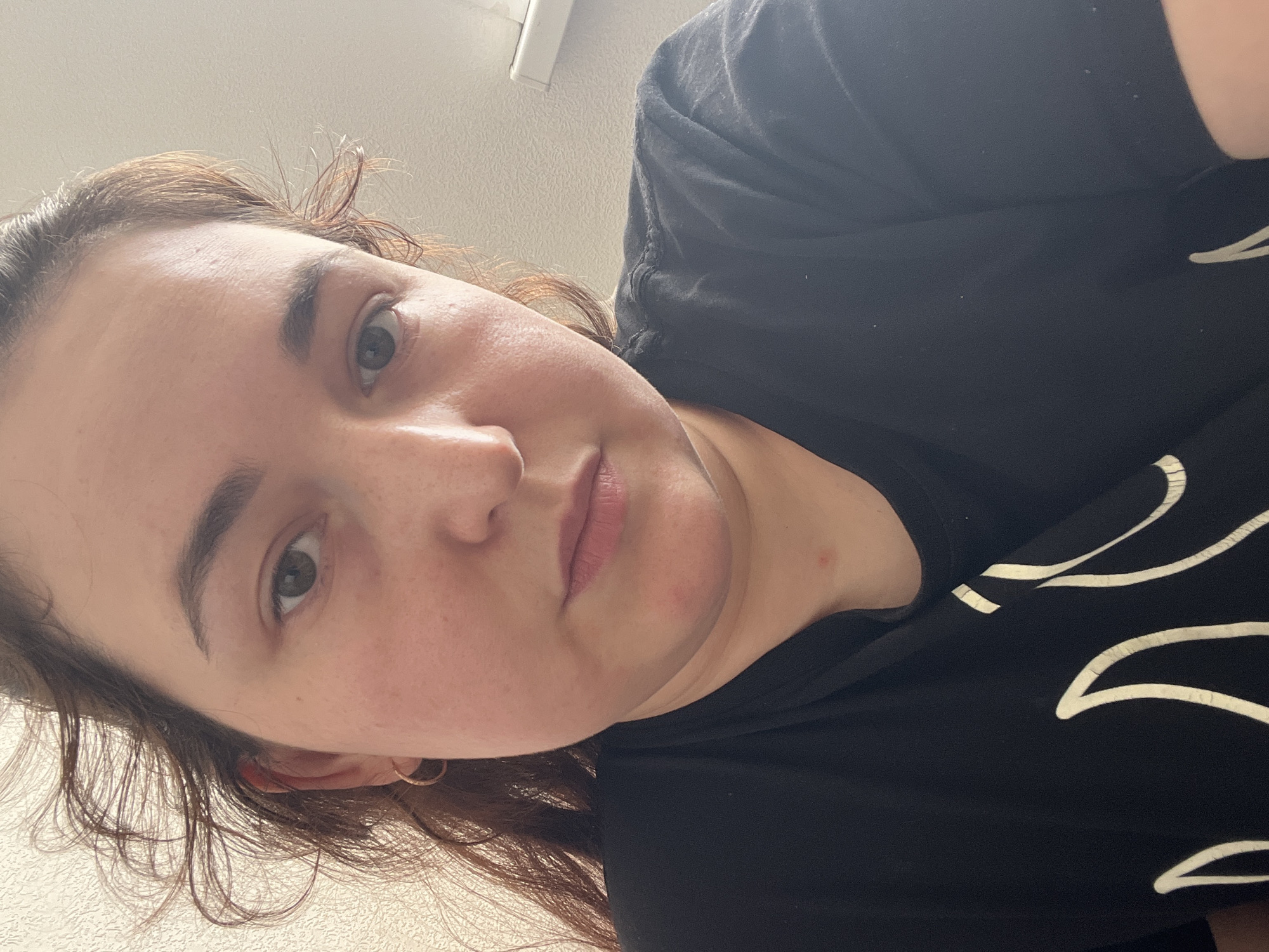The image is a close-up, frontal selfie of a young woman with fair skin, dark brown hair, and brown eyes. She is wearing a black t-shirt with a white flame-like graphic. Her hair is slightly windswept. The background is a textured, off-white wall and a white blind. There is a small red mark on her neck. The image provides no information about the location in Chelyabinsk, Russia.