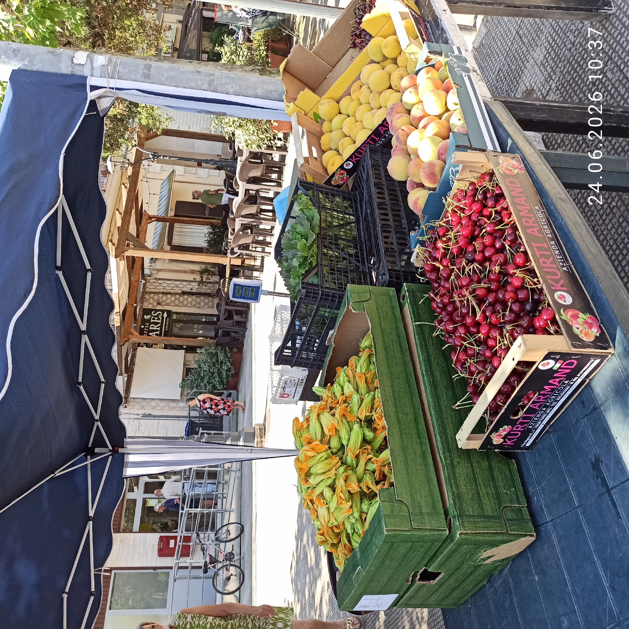 This is a horizontal, eye-level, outdoor, daytime photograph taken in Bitonto, Italy. The scene appears to be a fruit stand or market stall. The primary subjects are various fruits and vegetables displayed in crates and boxes. Prominently featured are bright red cherries, pale yellow peaches, and what appear to be zucchini blossoms. Some of the crates have labels with text. One label reads "KURTI ARMANDI AZIENDA AGRICOLA". Another label says "unifrutti". A digital scale is visible in the background.
In the background, there are glimpses of a building with windows and tables and chairs, suggesting an outdoor seating area, possibly for a cafe or restaurant. A dark blue awning provides shade over part of the fruit stand. There are also a few people visible, including a woman in a floral dress and a man in a green shirt. A bicycle is parked in the background. The presence of a scale, produce, and a canopy suggests a commercial setting. The date stamp "24.06.2026 10:37" indicates the time the photo was taken. The lighting suggests it is a sunny day.