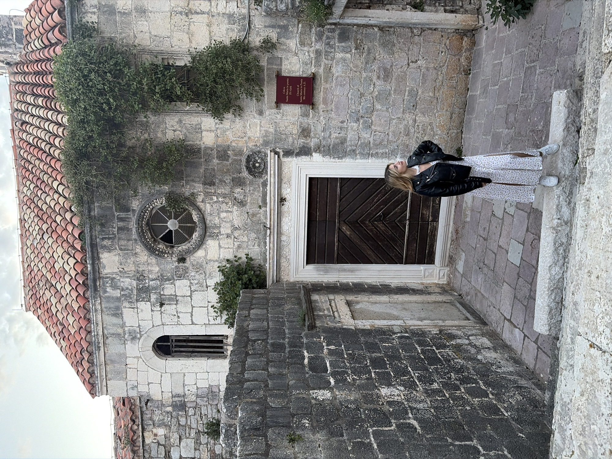 The image shows a building in the city of Budva, Montenegro, identified by a sign as the "Church of Santa Maria in Punta," dating back to the 9th century A.D. The church's facade is characterized by stone masonry, featuring a window with a round, decorative frame, and a wooden door with a chevron pattern. Patches of greenery grow on the stone walls.
A woman stands on stone steps in front of the building, wearing a white floral dress and a black leather jacket, looking up.