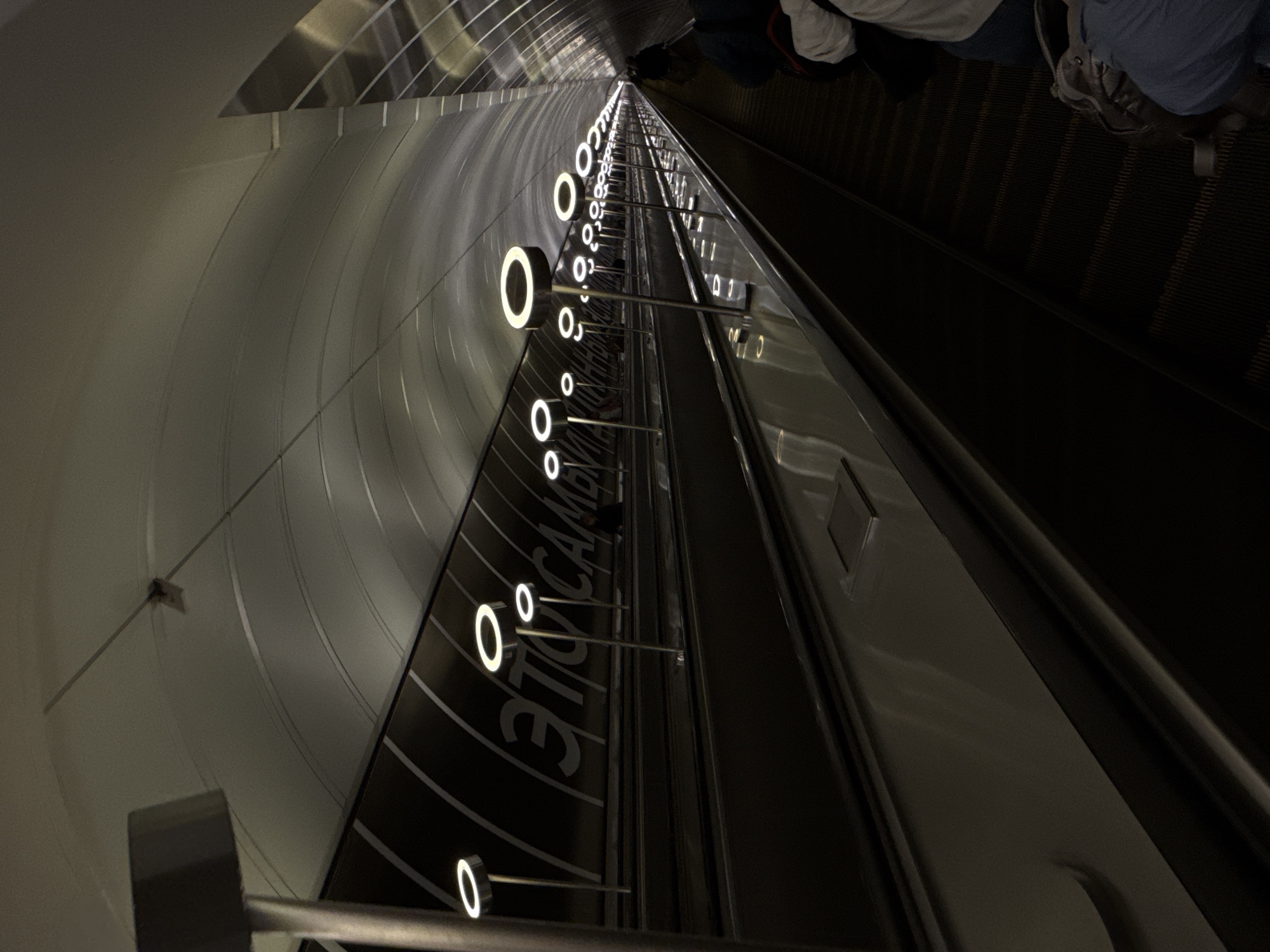 The image presents a long, deep escalator descending into what appears to be a modern subway or metro station. The perspective is taken from a high, tilted angle, looking down the length of the escalator, which emphasizes its considerable depth.
On the left side, the station's architecture features smooth, curved, light-colored metallic wall panels that create a tunnel-like environment. Parallel to the escalator, a prominent dark panel is adorned with a series of regularly spaced, glowing white circular lights, visually guiding the descent. Below these lights, Cyrillic text is visible, which clearly reads "ЭТО САМОЕ" (ETO SAMOYE).
Several people are visible on the upper right side of the escalator, primarily their legs and lower torsos, indicating they are descending. They are dressed in casual attire, with visible colors including blue, white, and darker tones. The escalator treads are dark and ribbed, contrasting with the lighter, reflective surfaces of the handrails and surrounding architectural elements.
The scene is illuminated by artificial lighting, with the bright circular lights casting reflections on the glossy surfaces, creating a sleek and somewhat futuristic atmosphere. The overall ambiance is dimly lit, highlighting the illuminated features. The architecture, deep escalator, and Cyrillic signage are strong indicators of a location like the Moscow Metro.