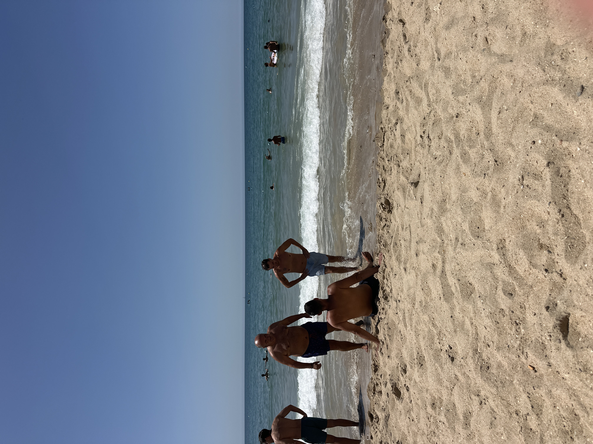 The image depicts a sunny day at a beach in Chiclana de la Frontera, Spain. The scene captures several men on the sandy shore and in the shallow waters of the ocean. The sky is clear and blue, with the sea stretching out to the horizon. In the foreground, a man is sitting on the sand, facing away from the camera. Behind him, four other men are standing near the water's edge. One man, slightly larger than the others, is in the center, appearing to converse with the man to his left. Another man stands to the right, with his hands on his hips. Further out in the ocean, several other individuals can be seen swimming and wading, and some are on a float. The waves are gentle, lapping at the shore. The overall atmosphere suggests a relaxed day at the beach.