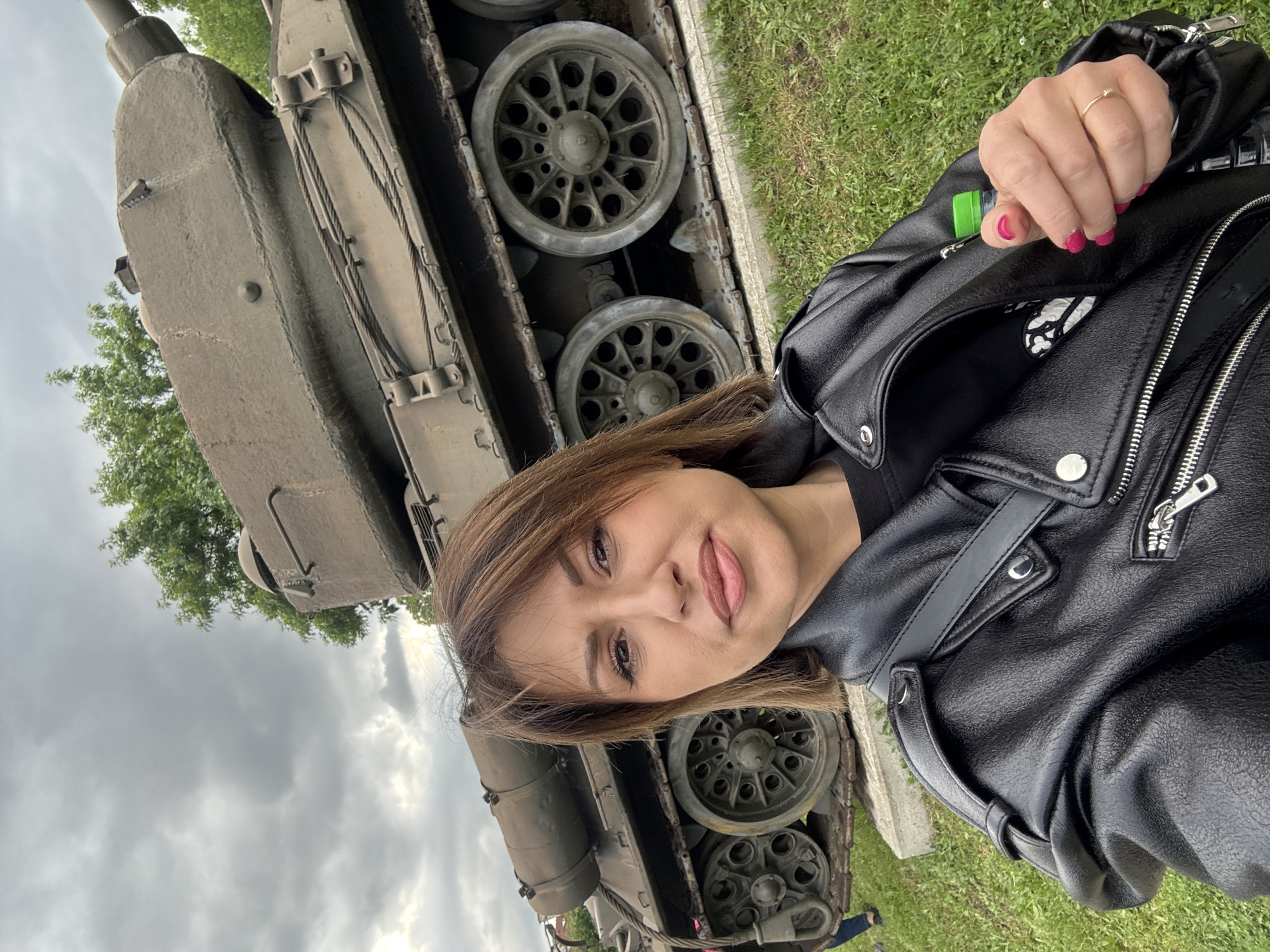 A woman is posing for a selfie in front of a tank. The tank is a military vehicle with visible wheels and tracks. The woman is wearing a black leather jacket. The sky is overcast with grey clouds. Green grass is visible near the tank. The location appears to be an outdoor display, possibly in a park or museum. This is likely located in Sofia, Bulgaria, given the request.