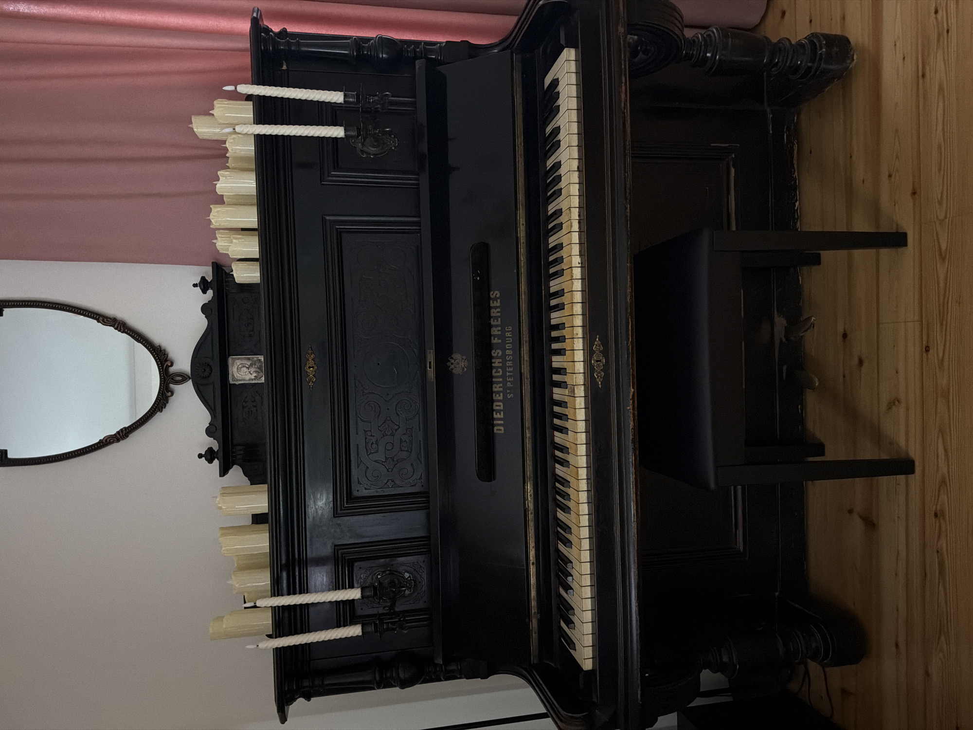 An antique black upright piano is positioned on a wooden floor, in front of a pink curtain and a wall. On top of the piano, near the back, are numerous unlit, cream-colored faux candles of varying heights arranged in a cluster. Two taller, cream-colored faux candles in dark candleholders are placed on the upper front of the piano. In the center of the piano's upper facade, there is a decorative plaque with the inscription "DIEDERICH FRERES" and below it, "ST PETERSBOURG". The piano keys are visible, with some yellowing on the white keys. A dark, square piano bench with four legs sits directly in front of the piano. Above the piano, a dark-toned ornate mirror with a curved top is mounted on the wall. The scene is lit by ambient light, creating shadows. There are no people or animals present. The location is not specified as Kolomna, Russia.