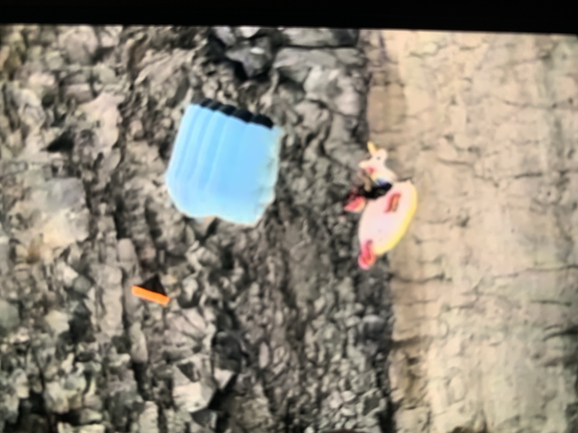 The image shows a blurred scene with a rocky background and a lighter-colored, textured foreground. In the upper portion of the image, an orange object, possibly a traffic cone fragment or marker, rests on the rocks. To the right of the orange object is a folded blue object, resembling a parachute or a deflated inflatable. In the lower part of the image, a pink inflatable ring with a unicorn design is visible, with a person or figure partially obscured on top of it. The overall impression is that of an outdoor scene, possibly a natural or semi-natural environment. There is no specific information to identify the location as Stavropol, Russia.
