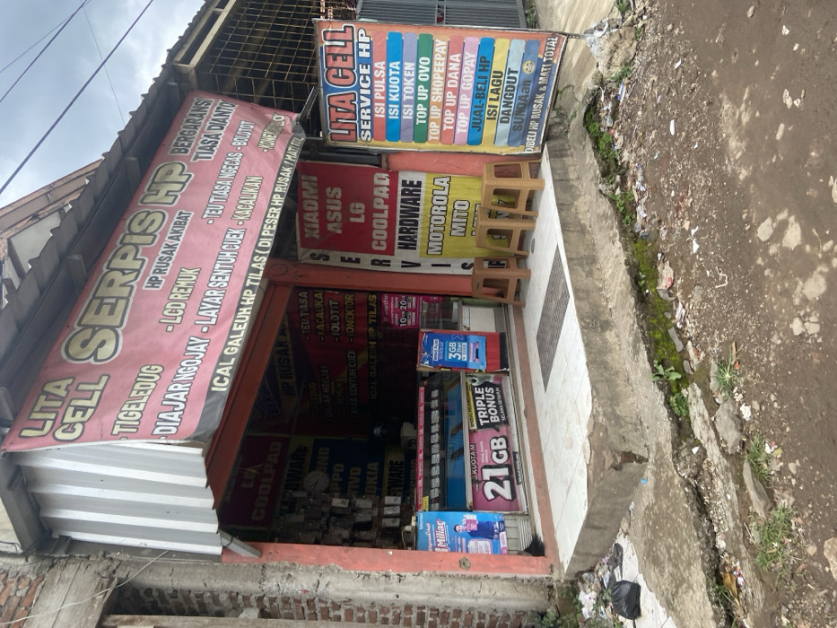 The exterior of a small storefront, located at latitude -7.021 and longitude 107.675, in the city of Baleendah, Indonesia, is visible. The shop's name is "Lita Cell," and it appears to be a cell phone repair and accessory shop. The storefront is decorated with colorful signage advertising the various brands and services offered, including "Serpis HP," "Lita Cell Service HP," and "Xiaomi, Asus, LG, Coolpad, Hardware, Motorola, Mito." The store is decorated with a red, white, and yellow color scheme, with various brands of cell phone cases and accessories visible in the window display. Two plastic chairs sit in front of the store, indicating the presence of people waiting to be served. The shop front is set back from the sidewalk, with an open concrete area in front, which is unkempt, and littered with trash. There is no one currently in the immediate area of the store. There is a plastic bag of trash on the ground next to the storefront, and various pieces of litter are scattered on the sidewalk. There is green moss growing in the cracks of the concrete sidewalk. The weather appears to be overcast. The storefront is built in a similar style to the other buildings in the area, with a concrete facade and a simple design. There is a visible power line running across the top of the image, which appears to be above the storefront and behind the signage. The storefront appears to be a small, local business that provides services and products for the surrounding community. The scene is a typical example of a small, local shop in Indonesia.