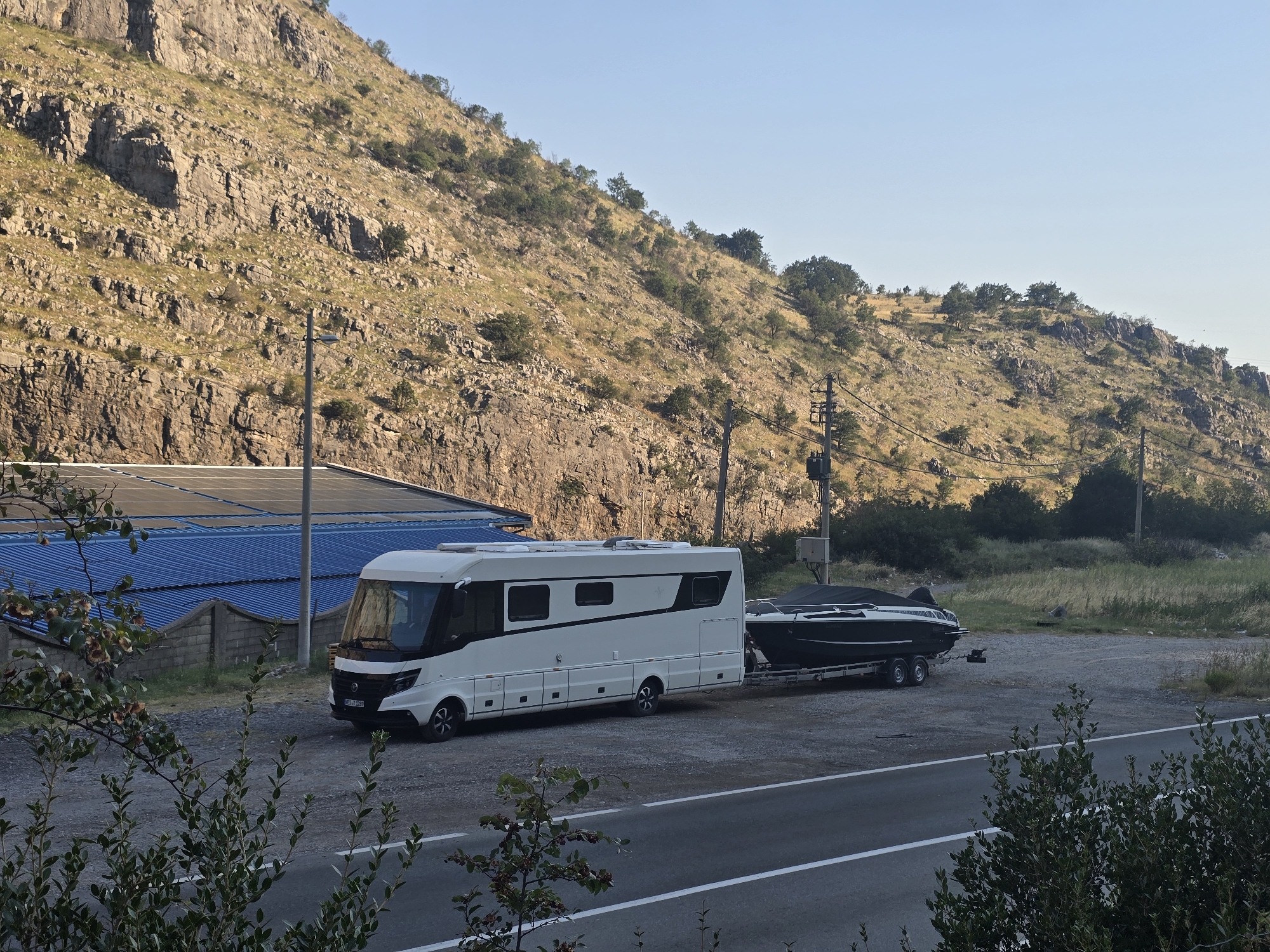 The image depicts a white RV and a black boat on a trailer parked in a gravel lot next to a paved road. The RV is a large motorhome with multiple windows and black accents. The boat is sleek and modern, covered with a dark tarp. Behind the vehicles, there is a building with a blue roof and a stone wall. The background features a steep, rocky hillside covered with sparse vegetation and trees, with a clear blue sky above. Power lines and poles are visible on the hillside. The scene appears to be set in a rural or semi-rural area, possibly on the outskirts of a city like Podgorica, Montenegro, given the hilly terrain and the presence of infrastructure. There are no people visible in the image.