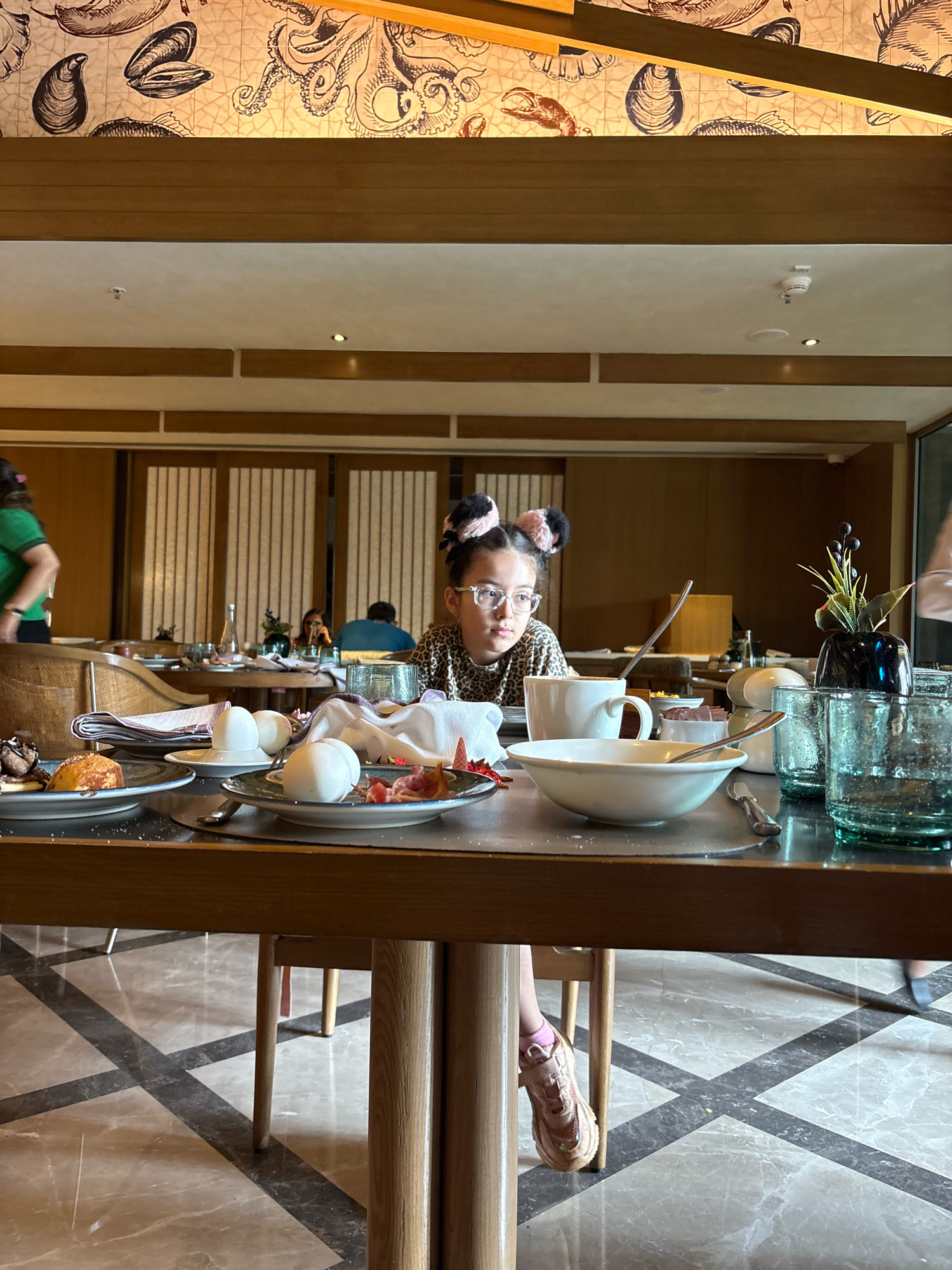In the city of Vagator, India, a young girl with eyeglasses and fluffy pink and black hair accessories sits at a wooden table in a well-lit restaurant. She is wearing a leopard-print top. The table is laden with breakfast items, including plates with food, bowls, cups, and glasses. One plate in the foreground holds what appears to be a pastry and some chocolate sauce, while another has white boiled eggs. A white bowl with a spoon sits in the center of the table, next to a white mug. To the right, there are two green glass tumblers and a small decorative vase with a plant. The background reveals other diners and decorative wooden panels on the walls, with an overhead section featuring a mural of various sea creatures. The floor is tiled with a geometric pattern of dark gray and light marble. The overall impression is of a casual family meal in an upscale dining establishment.