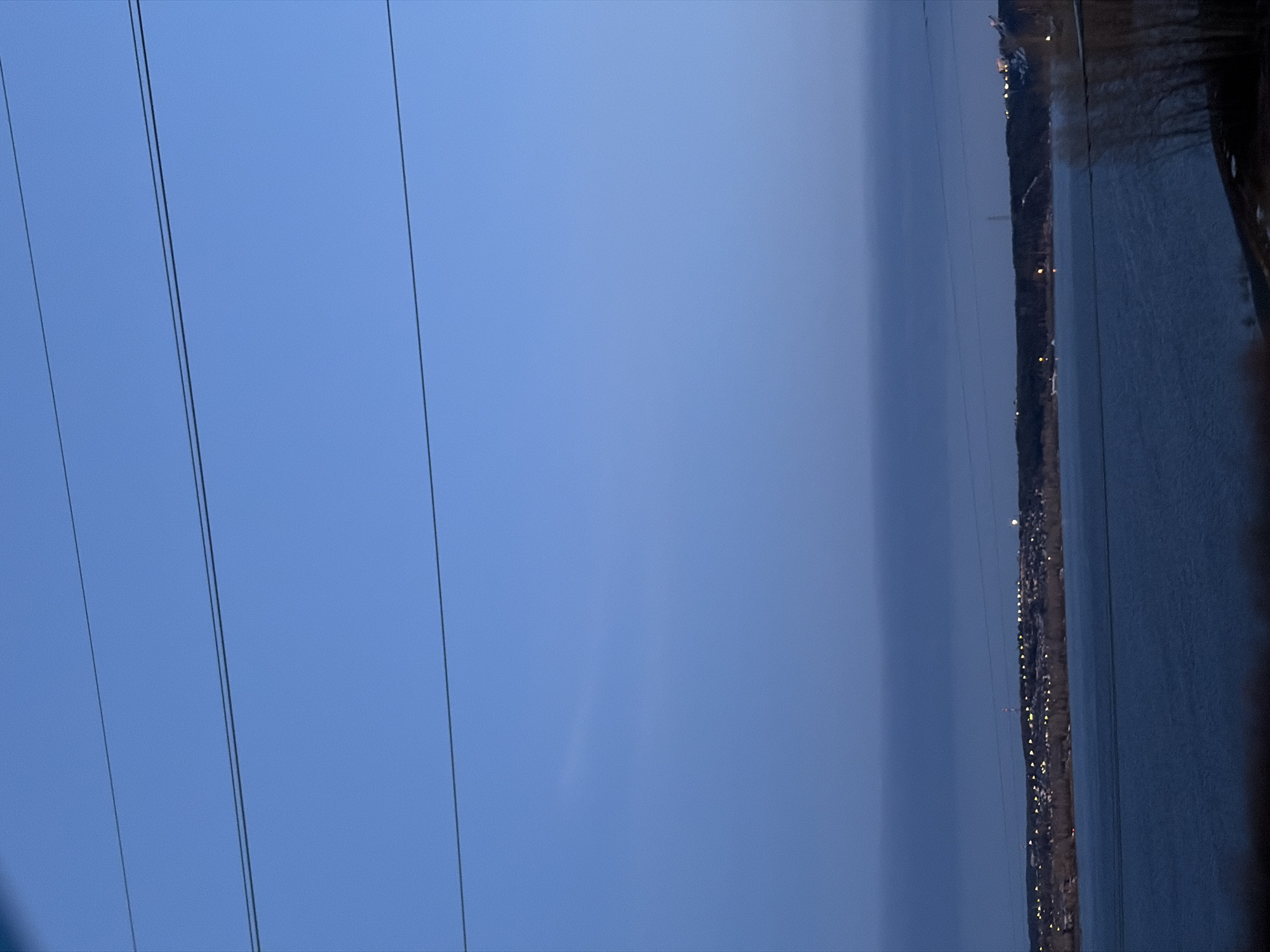 The image shows a body of water on the right side, with a shoreline densely lit by many small lights, likely a city or town at night. The image appears to be taken at dusk or dawn. The sky is a gradient of blue, with some power lines stretched across the left side of the frame. The location is likely Chaikovsky, Russia, based on the geographical features and urban development present.