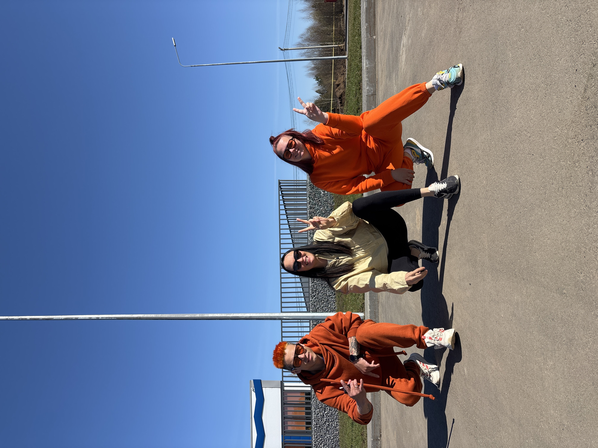 Three individuals pose in an outdoor setting in Votkinsk, Russia. The location appears to be a paved area near a fence and a building with blue and white accents. The sky is clear and blue. The individuals are squatting in a vertical line, each facing the camera and gesturing with a "peace" sign. The person at the top is wearing an orange tracksuit and sunglasses. The middle person is wearing a tan jacket and black pants with sunglasses. The bottom individual is wearing an orange tracksuit, sunglasses, and has orange hair. They are also holding a stick-like object.