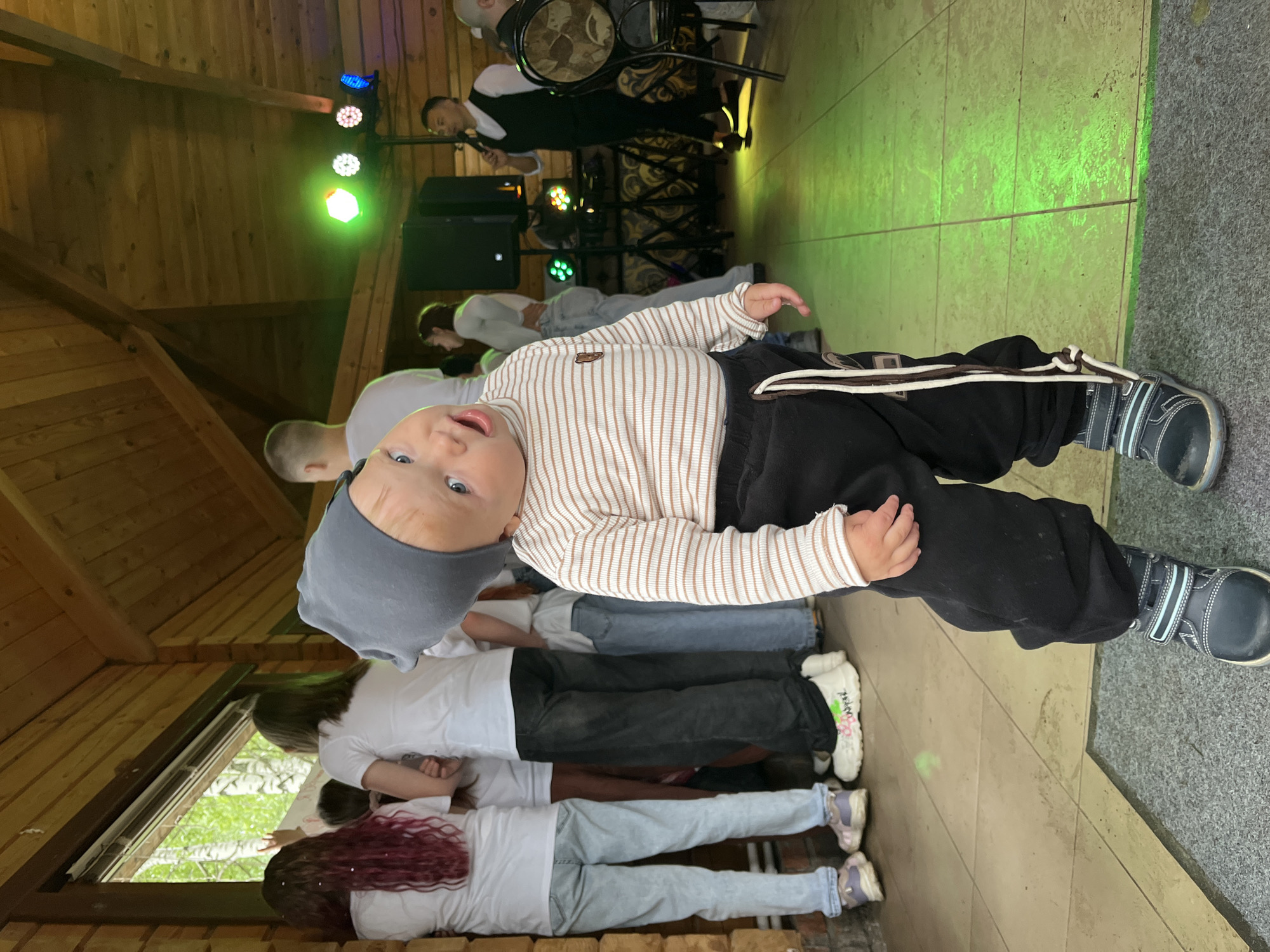 A baby wearing a striped shirt and gray hat lies on the floor, surrounded by several people who are standing. The baby is also wearing black pants with a white rope tie. The scene appears to be indoors, with wooden walls and ceiling, and green stage lighting in the background, suggesting an event space or venue. A man is visible on a stage, singing into a microphone. The floor is tiled in a grid pattern. The location is the city of Chaikovsky, Russia.
