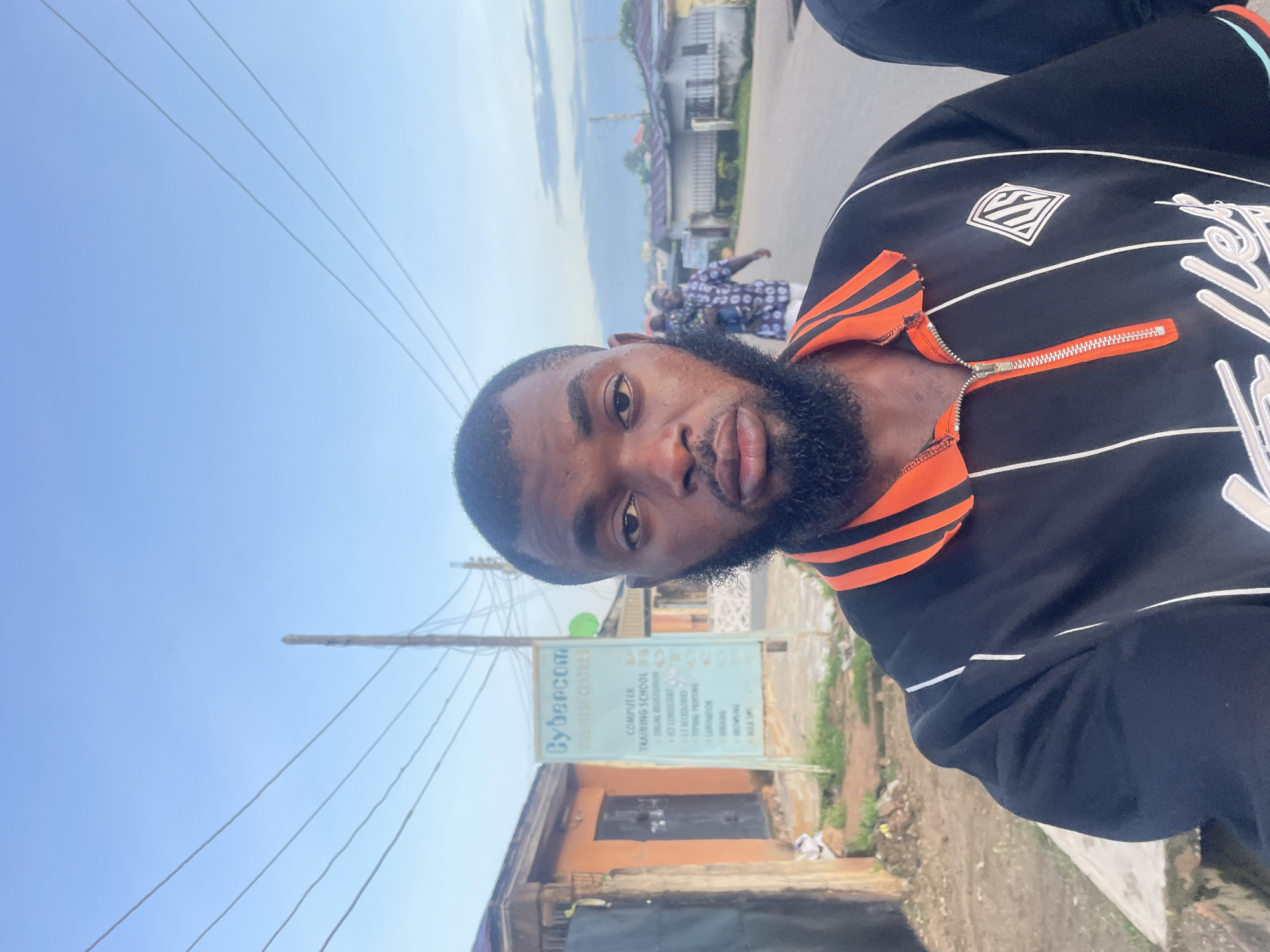 A head and shoulders selfie of a Black man with a beard is taken outdoors in Jos, Nigeria. He is wearing a black and orange zippered polo shirt. Behind him, a sign for "Cybercom Business Centre" is visible, listing services like "Computer Training School" and "ICT Consultant." A street with houses and power lines stretches into the distance, under a pale blue sky with scattered clouds. Another person carrying a child is walking away in the background.