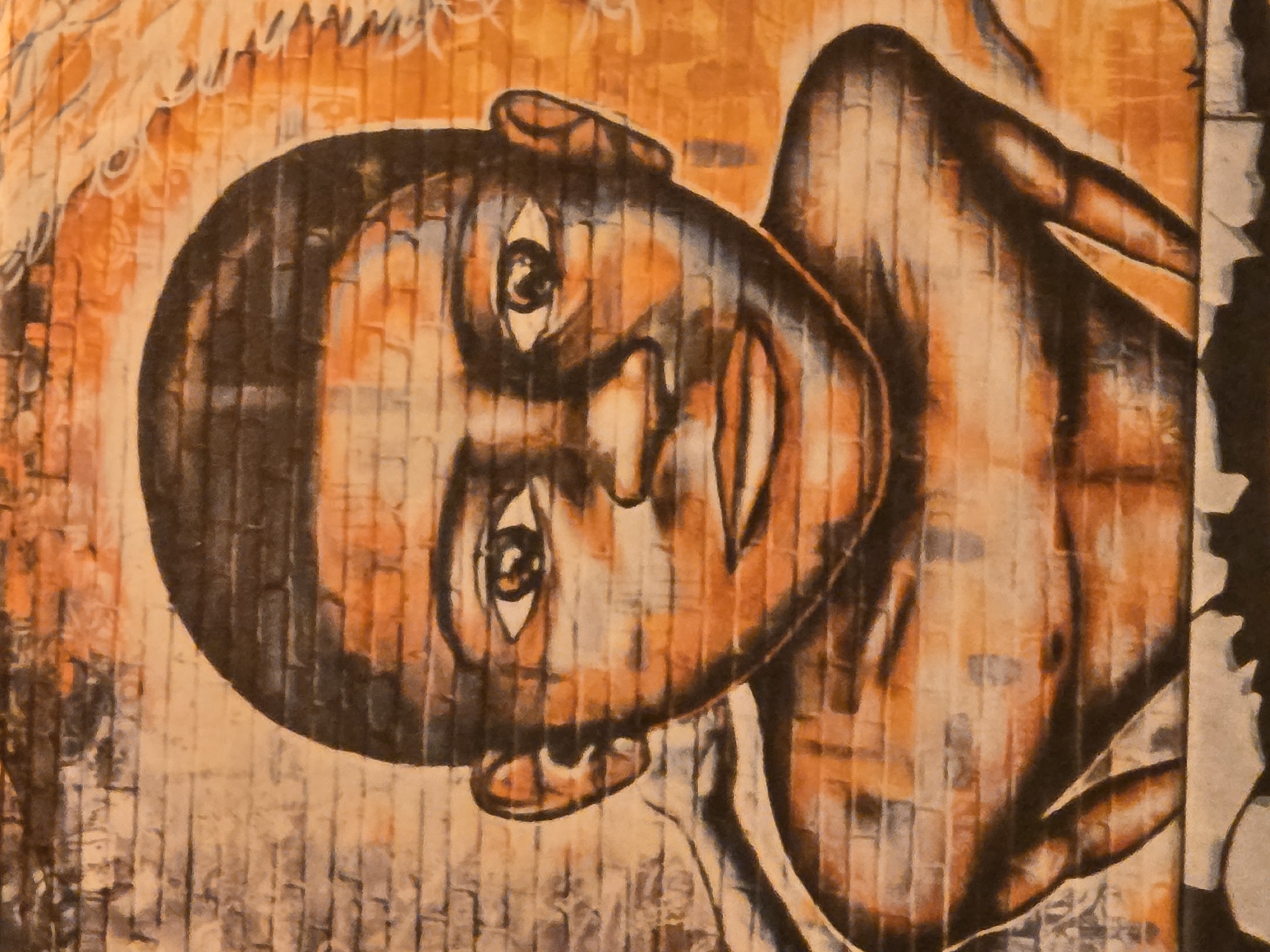 The image is a close-up of a mural depicting a person's head and torso. The subject has dark skin and short hair. The mural is painted with an orange and brown color palette. The background texture appears to be brick, suggesting the mural is on a brick wall. The location is in an unknown city and region.