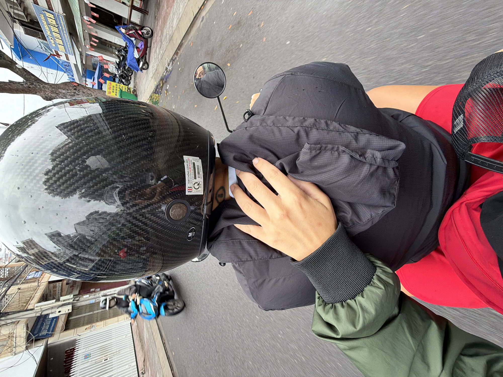 The image is a point-of-view shot from the back of a motorcycle in Nha Trang, Vietnam. The rider, wearing a black carbon fiber helmet with water droplets, is being followed by a person in a green jacket. The passenger's right hand is resting on the rider's black jacket. The passenger is also wearing red shorts and a black mesh hat. The motorcycle is on a wet asphalt road, suggesting it may have rained recently. Buildings with signs in Vietnamese line the street. A blue scooter is visible in the distance on the left, and several other motorcycles are parked on the right side of the street.