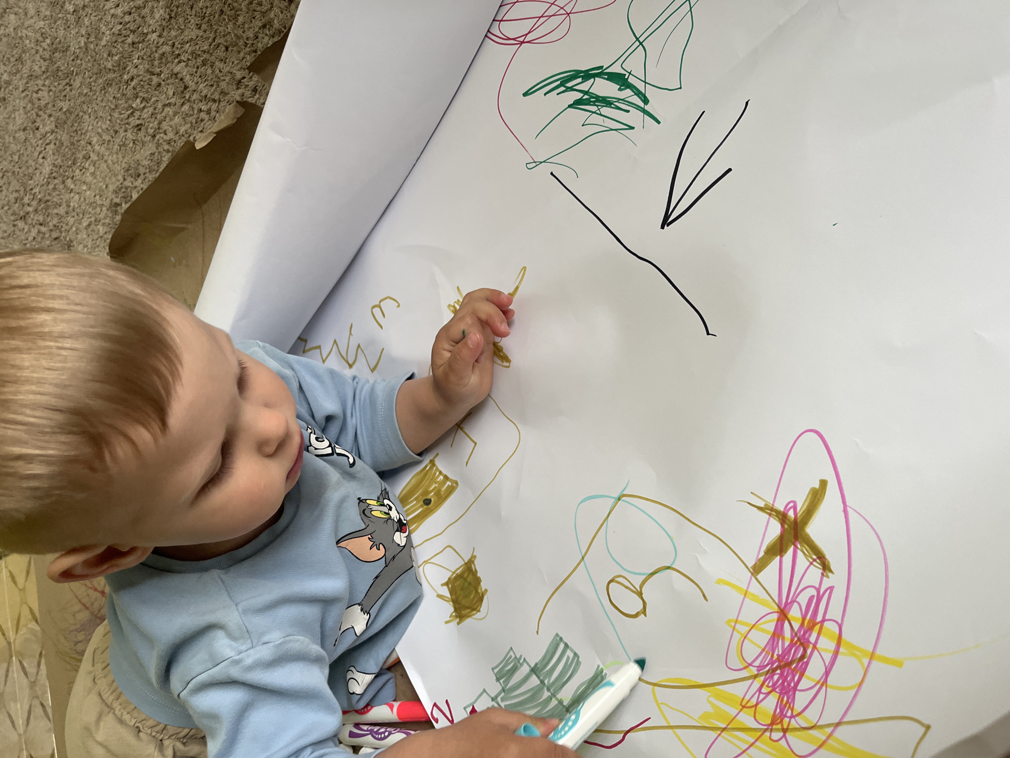 A blond-haired toddler wearing a light blue long-sleeved shirt with a Tom and Jerry graphic is drawing on a large sheet of white paper. The child is focused on the paper, with their head bowed. Their right hand is holding a green marker, and their left hand is visible near the paper. The paper is covered in various scribbled lines and shapes drawn in multiple colors, including yellow, pink, brown, green, and blue. Some of the drawings appear to be abstract scribbles, while others suggest rudimentary shapes like squares or crosses. A large roll of white paper is partially visible in the background, along with a cardboard box and carpeted floor. The image appears to be taken indoors, likely in a home environment, with no discernible connection to the city of Onokivtsi, Ukraine.