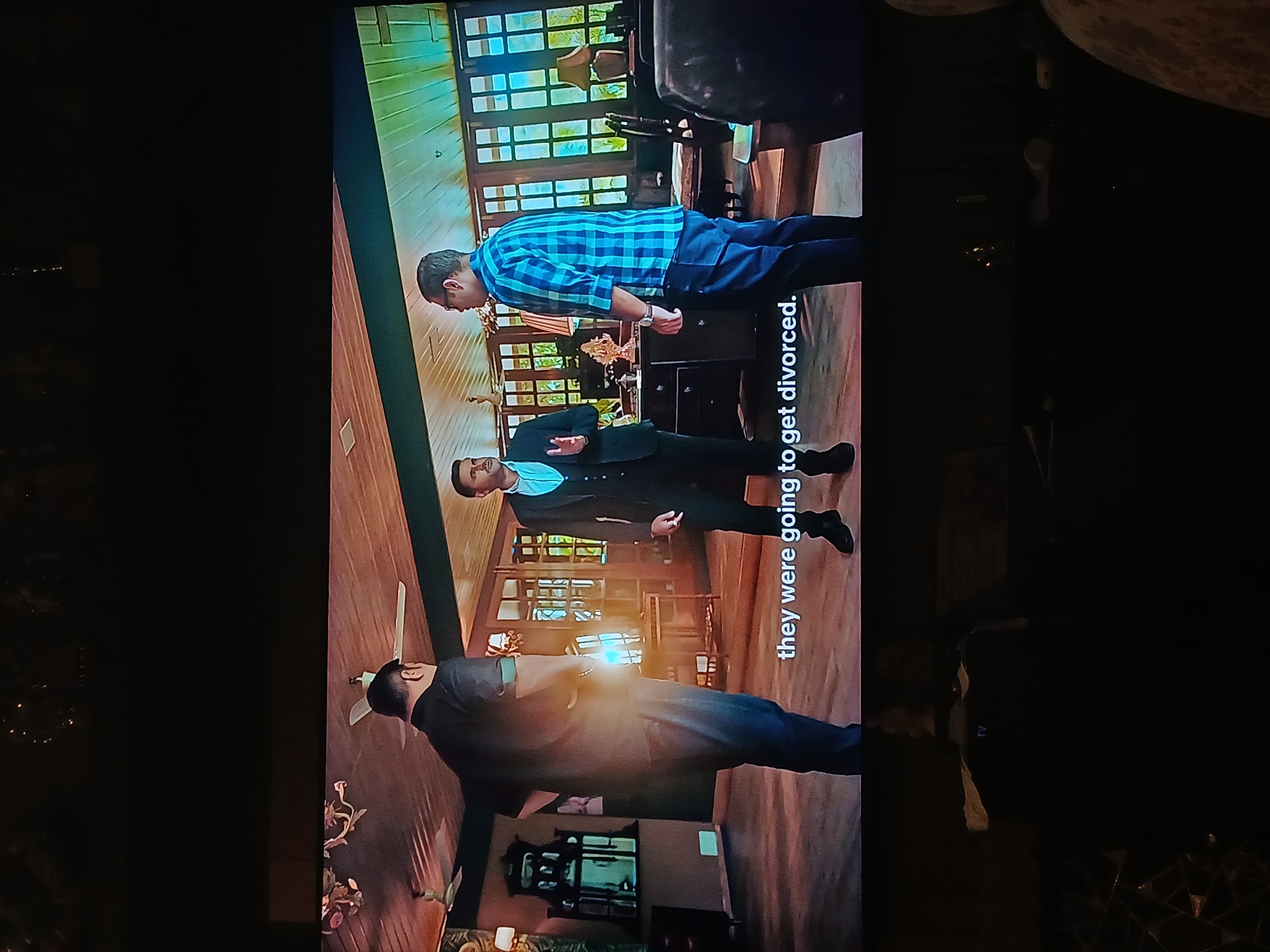 The image shows a television screen displaying a scene from a movie or show. Three men are visible in the scene, standing in what appears to be a wooden-floored room with multiple windows. One man is wearing a blue and white checkered shirt and dark pants. Another man is in a dark suit with a light-colored shirt. The third man is also wearing a dark suit and glasses. Text overlay on the screen reads "they were going to get divorced." The overall image is taken in a dark room, with some surrounding objects visible, including what appears to be a marble surface. There is no visual information indicating the location is in Bengaluru, India.