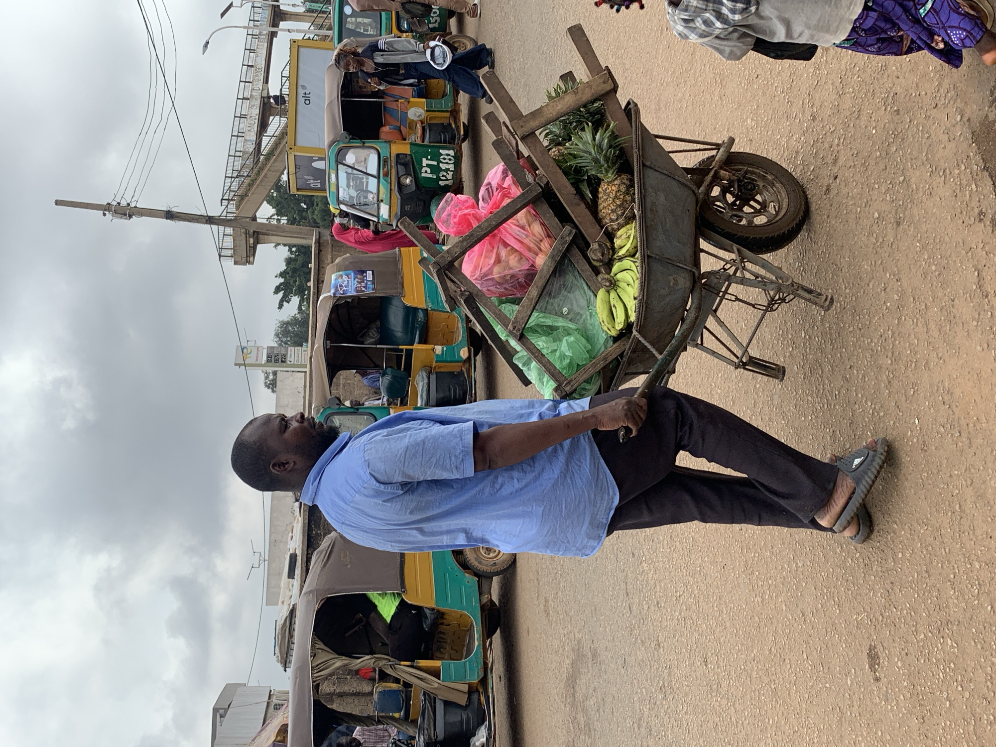 An overcast day in Gura Top, Nigeria, finds a man pushing a wheelbarrow laden with produce and other items. The wheelbarrow is filled with bananas, pineapples, and what appear to be some pink-bagged goods, possibly more fruit or vegetables. Several wooden planks are precariously stacked on top of the load. The man, wearing a blue button-down shirt and dark trousers, is seen from the side, his face turned away from the camera. He wears dark sandals.
In the background, several auto-rickshaws, common in Nigerian cities, are parked or in motion. One has the license plate "PT 12181". A pedestrian walkway or bridge is visible in the distance, along with utility poles and power lines. A young child, wearing a plaid shirt and patterned purple skirt, is partially visible on the right side of the frame, their feet facing the viewer. The ground is a rough, sandy texture, typical of unpaved urban areas. The overall scene depicts a busy street or marketplace environment.