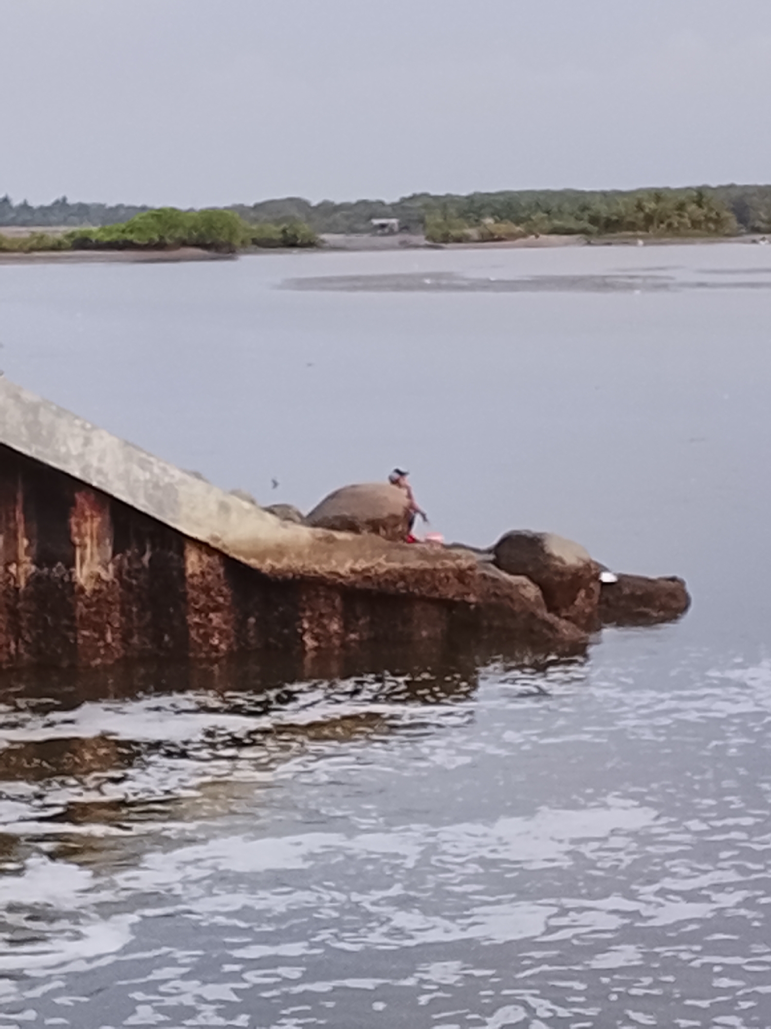 A person is sitting on rocks at the edge of a body of water. The person appears to be male and is wearing a dark shirt and red shorts. They are holding a fishing rod and a bucket. The rocks are part of a concrete structure that is partially submerged in the water, likely a pier or breakwater. The water is calm with some ripples and foam. In the background, there is a shoreline with trees and vegetation. The sky is overcast and gray. The scene is likely located in Lhokseumawe, Indonesia, which is a coastal city known for its fishing industry. The overall impression is one of a peaceful, everyday scene in a coastal environment.