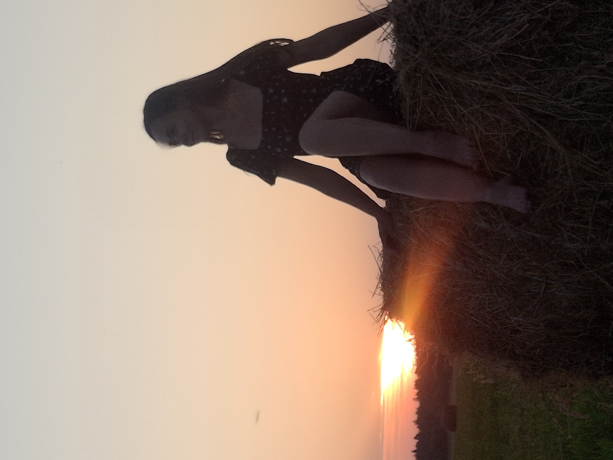 A silhouette of a young woman in a dark floral dress sits on a large hay bale in a field at sunset. The sky is a gradient of pale yellow and pink. The sun is low on the horizon, casting a warm glow over the scene. The woman is looking to her left, with her legs crossed. The hay bale is rough and textured, with strands of hay visible. In the background, there is a dark line of trees and another hay bale in the distance. The overall impression is one of peace and tranquility. The location is the outskirts of Perevoznoe, Russia.