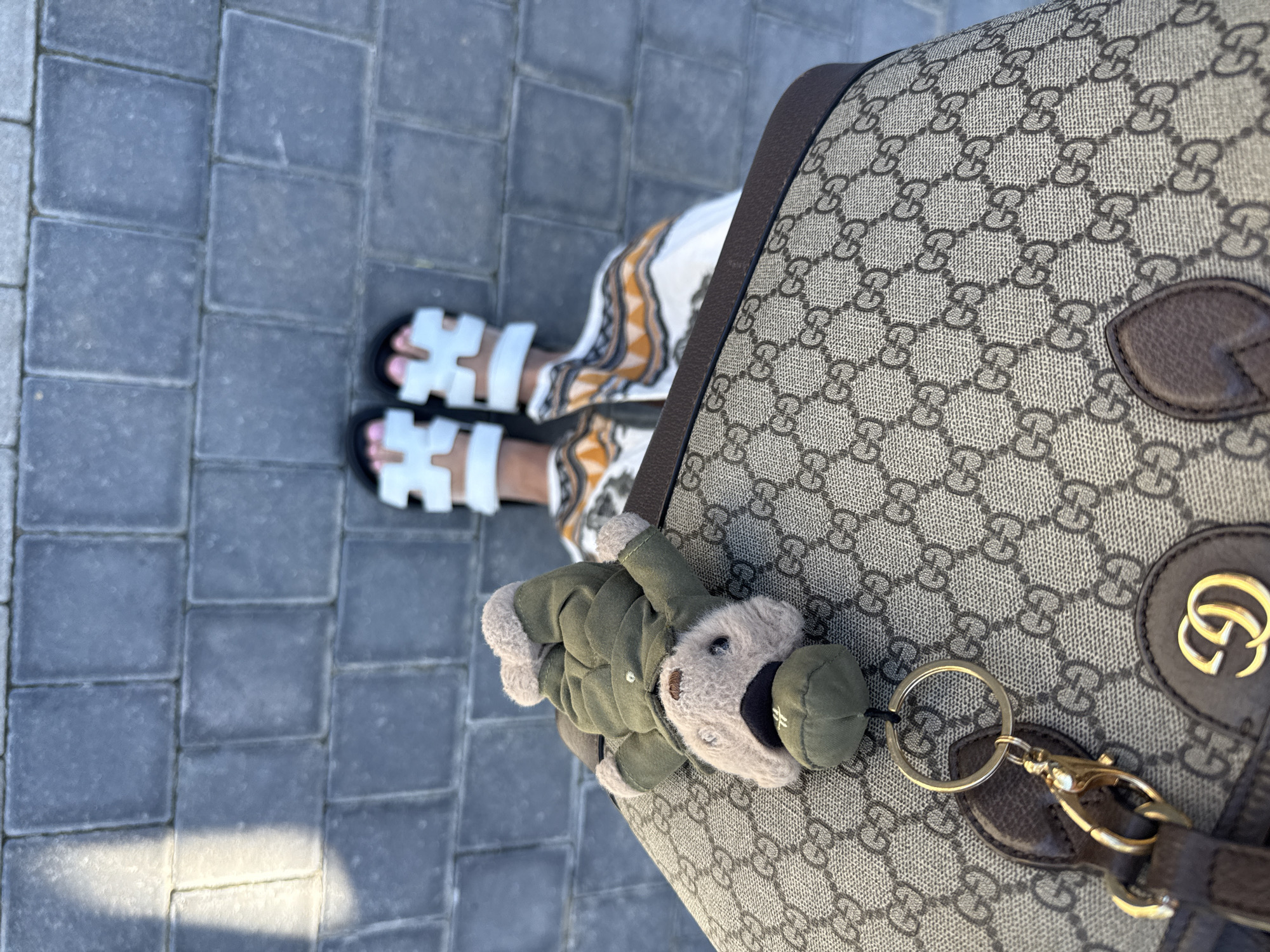 The image shows a person standing on a gray brick sidewalk, likely in an urban area. The person is wearing white sandals with black soles and a white dress with an orange and brown pattern. A Gucci bag with the classic "GG" logo is visible, adorned with a teddy bear keychain wearing a green jacket and cap. No specific landmarks or features clearly indicate the location as Cape Town, South Africa.