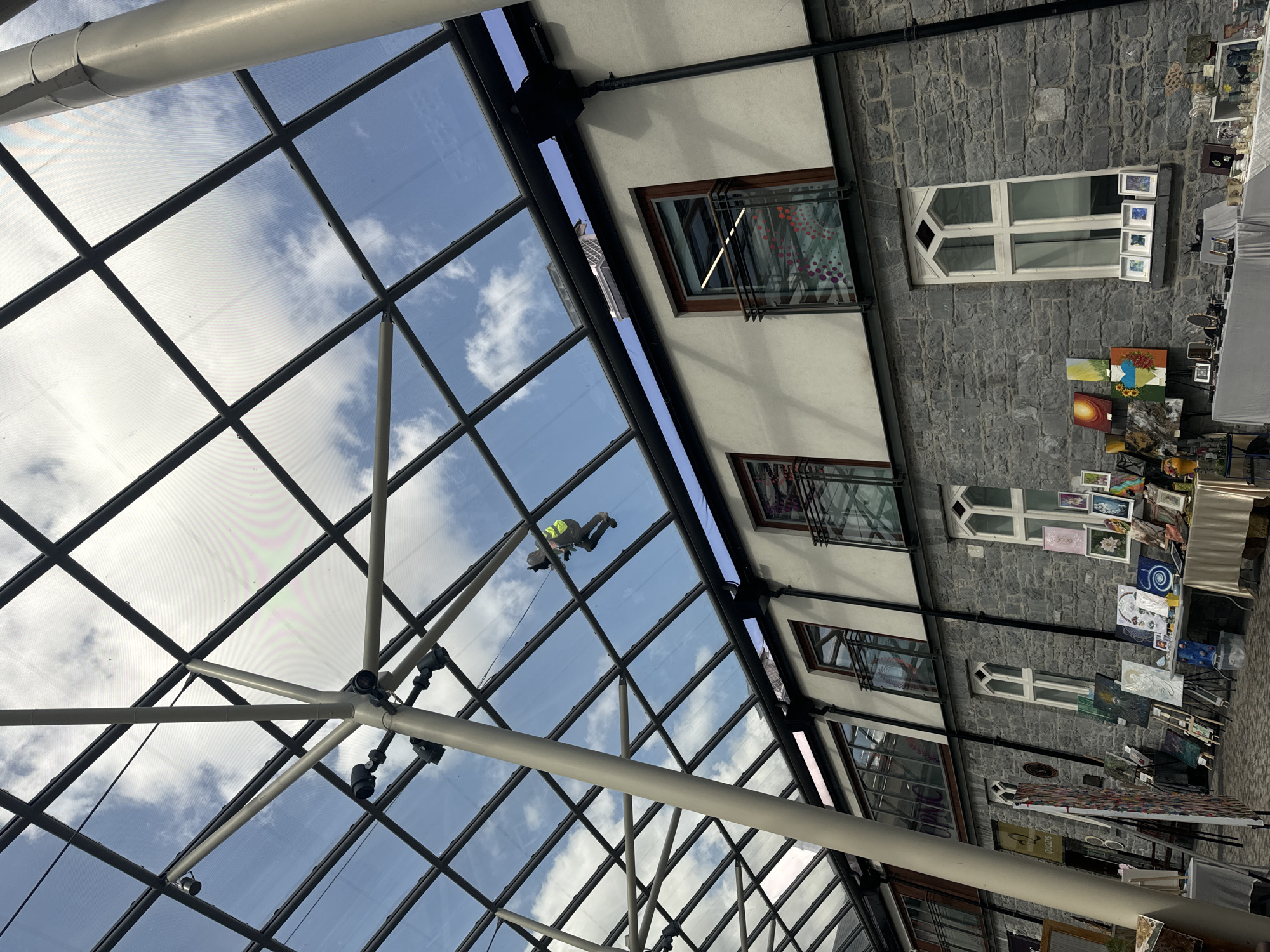 The image captures a view looking up at a glass-paned, lattice-like ceiling supported by a central pillar. Through the ceiling, a blue sky with scattered white clouds is visible. A person in a yellow safety vest and dark clothing is suspended from a cable system, seemingly cleaning or inspecting the ceiling. Below the ceiling, a three-story building with stone and white-painted facades is visible. The building features multiple windows, some with balconies. The ground level appears to be an indoor market or exhibition space, with various artworks displayed on easels and tables, including paintings and framed pieces. This scene is likely located within the Kilkenny Design Centre in Kilkenny, Ireland, given its architecture and the context of an art exhibition.