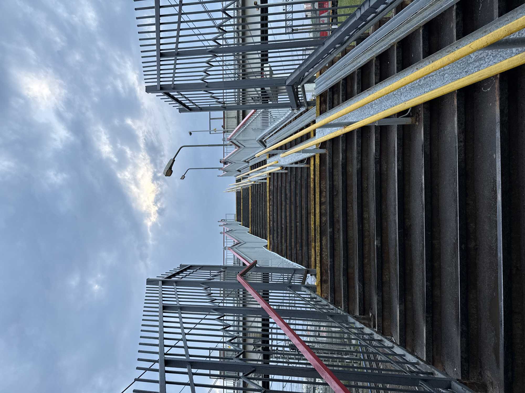 The image depicts a staircase leading up to a pedestrian overpass, likely in an urban area. The stairs appear to be constructed of dark metal, with yellow and red handrails. Metal fencing lines both sides of the staircase. The sky is overcast with cloudy formations. Based on the architectural style and the presence of overhead power lines, the location is possibly in Moscow, Russia, although specific landmarks aren't visible to confirm this. There are no people visible in the image.