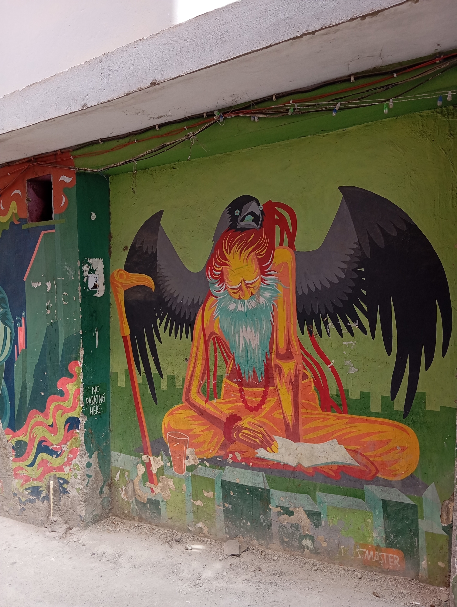 The image shows a mural painted on a wall in Jonk, India. The mural depicts a seated figure with a long white and turquoise beard and orange skin, wearing a string of prayer beads. The figure is meditating, holding a book or paper in his lap, with a walking stick leaning to his left. Behind the figure is a large, black bird, possibly a raven, its wings spread wide. To the left of the figure is a cup and a partially damaged wall with another mural. The mural is signed "ST. MASTER". Above the mural, wires and electrical components are visible. The wall shows signs of wear and damage. The ground is a light gray concrete or dirt.