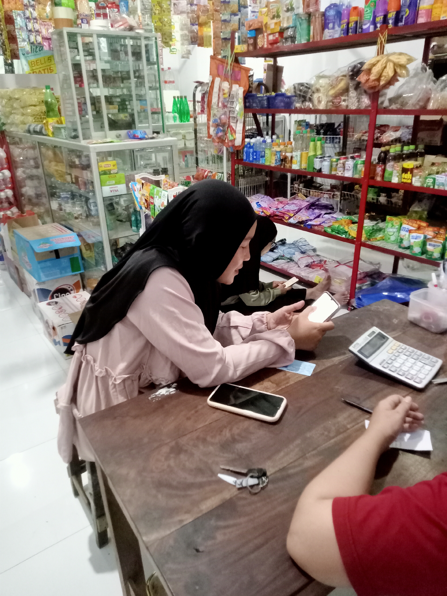 Two individuals are seated at a wooden table in what appears to be a small convenience store in Simo, Indonesia. The person in the foreground, wearing a pink long-sleeved top and a black hijab, is holding a smartphone. To their right, a calculator is placed on the table. In the background, shelves are stocked with various products, including food items and beverages, indicative of a retail environment. Another person is partially visible in the background, also looking at a phone. The overall setting suggests a casual interaction within a commercial space.