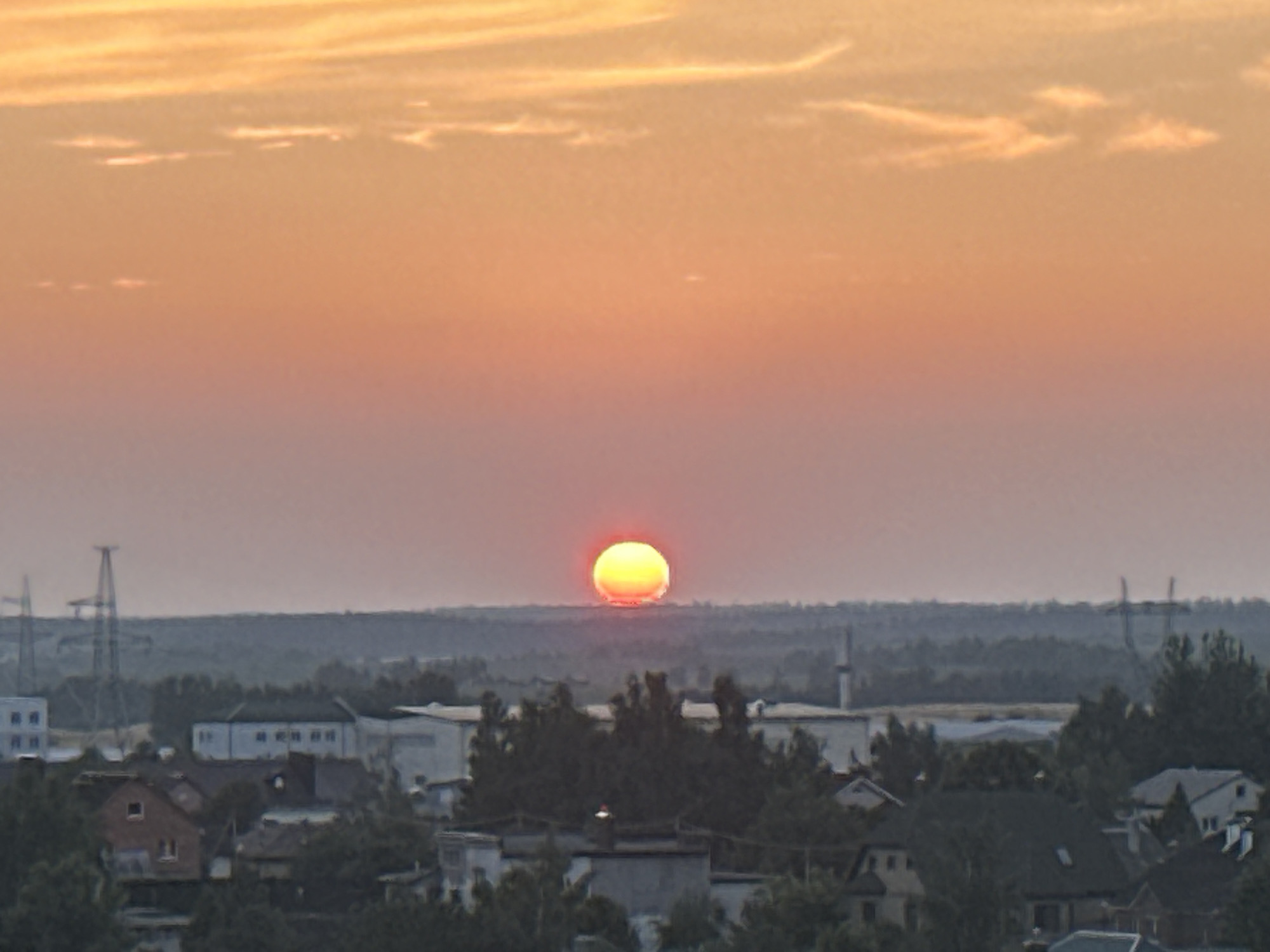 The image captures a sunset over a suburban landscape. The sun, a vibrant orange disk, is low on the horizon, partially obscured by a hazy atmosphere. The sky transitions from a bright orange at the top to a softer pink and then a pale blue near the horizon. Below the sky, a band of dark green trees stretches across the middle ground. In the foreground, a collection of houses and buildings with various rooflines and architectural styles are visible, interspersed with more trees. Power lines and transmission towers are visible in the distance, suggesting the presence of infrastructure within or near the settlement. The scene depicts a residential area at dusk, with the setting sun casting a warm glow over the landscape. This image is of the Шчомысліцкі сельскі Савет, Belarus.