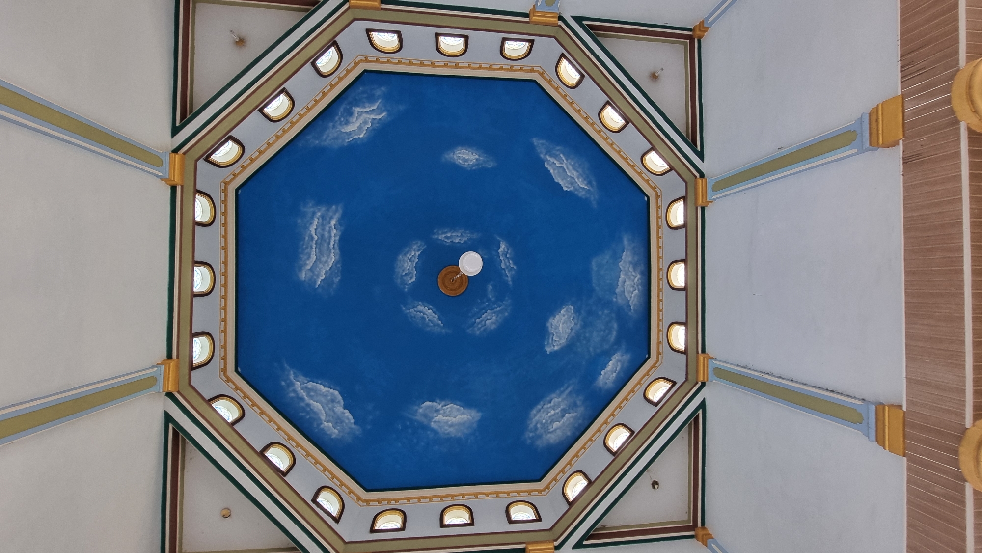 The image shows an octagonal ceiling decorated with a blue sky and white clouds mural. Along the edges of the ceiling, there are small arched windows with light coming through. The ceiling is framed by decorative borders in various colors including gold, green, and brown. In the center of the ceiling hangs a light fixture. The lower portion of the image reveals white walls supported by pillars with similar decorative elements. The overall style suggests an interior architectural space, possibly a building in Meunasahlhok, Indonesia, designed for aesthetic appeal.