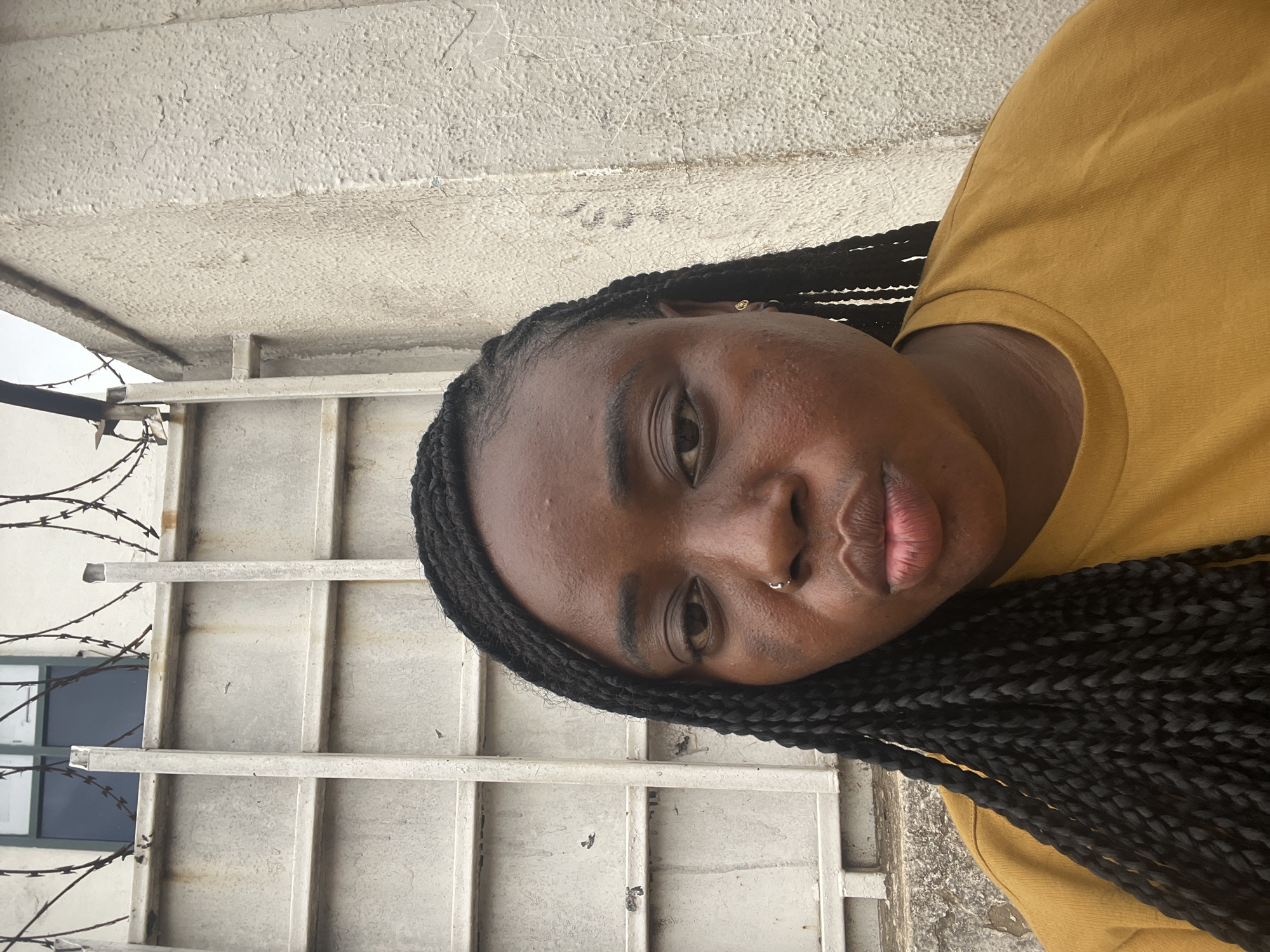A close-up portrait of a young Black woman is presented, rotated 90 degrees clockwise. She has dark skin, long dark braided hair, and wears a mustard yellow t-shirt. A small silver hoop pierces her septum, and a subtle gold stud is visible in her right ear. Her gaze is directed at the viewer, with a neutral expression and slightly parted, full lips.
The setting is outdoors in Abuja, Nigeria, under bright daytime conditions. The background consists of a textured, light-colored wall, likely concrete, showing some signs of wear and cracks. Behind her, a grey metal fence or gate with horizontal bars is partially visible. To the left, strands of barbed wire are wrapped around a vertical support, and a dark-paned window is glimpsed through the fence structure. No specific activity beyond posing for the camera is evident, and no visible text is present.
