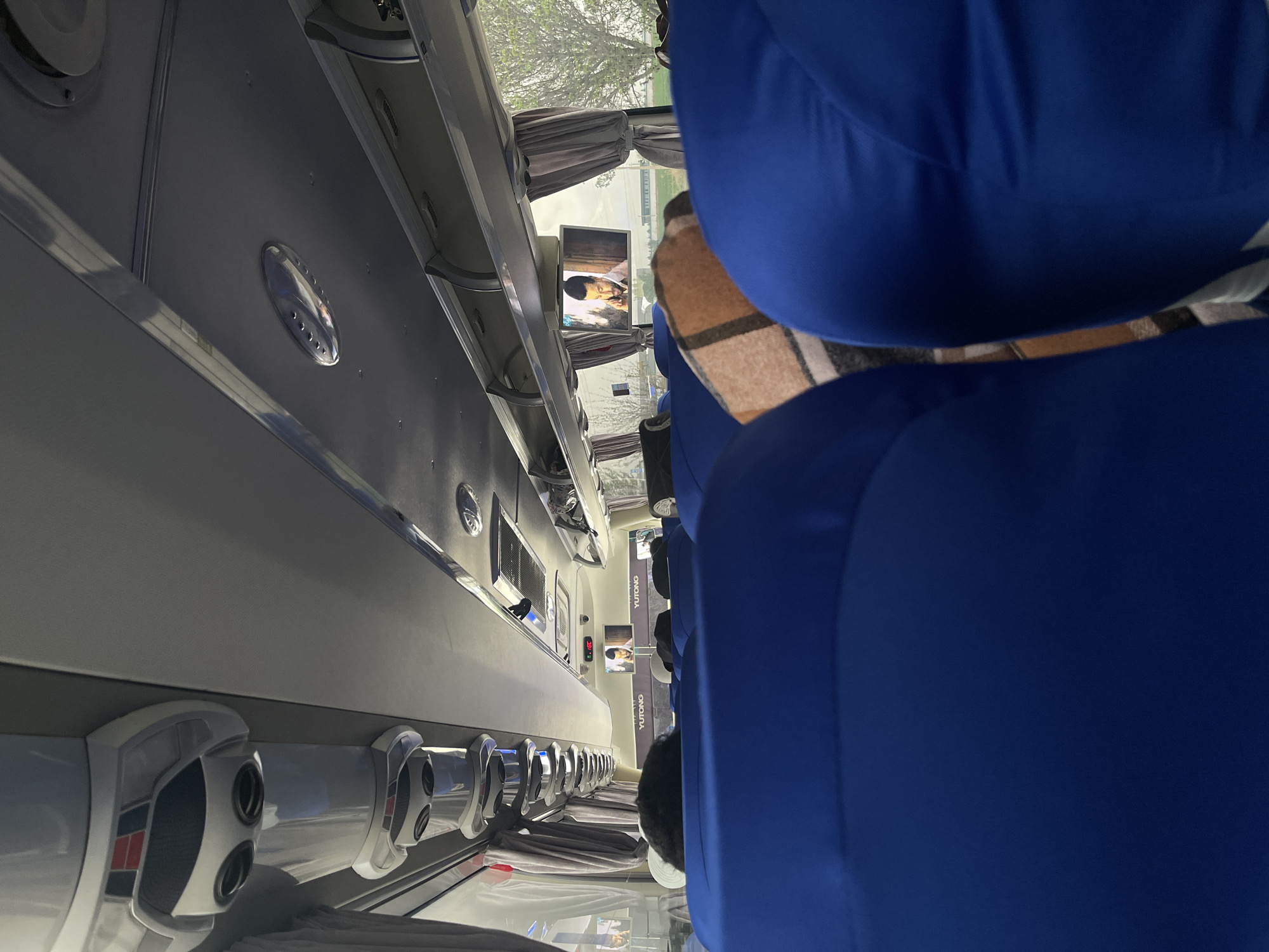The image depicts the interior of a Yutong passenger bus, likely traveling along a route in Karmana, Uzbekistan, based on the displayed name. The bus features blue upholstered seating and individual entertainment screens mounted above each row. The lighting fixtures and overhead storage compartments are visible. Some passengers are seated, with hats and blankets. The exterior is seen through the windows, where trees are visible.
