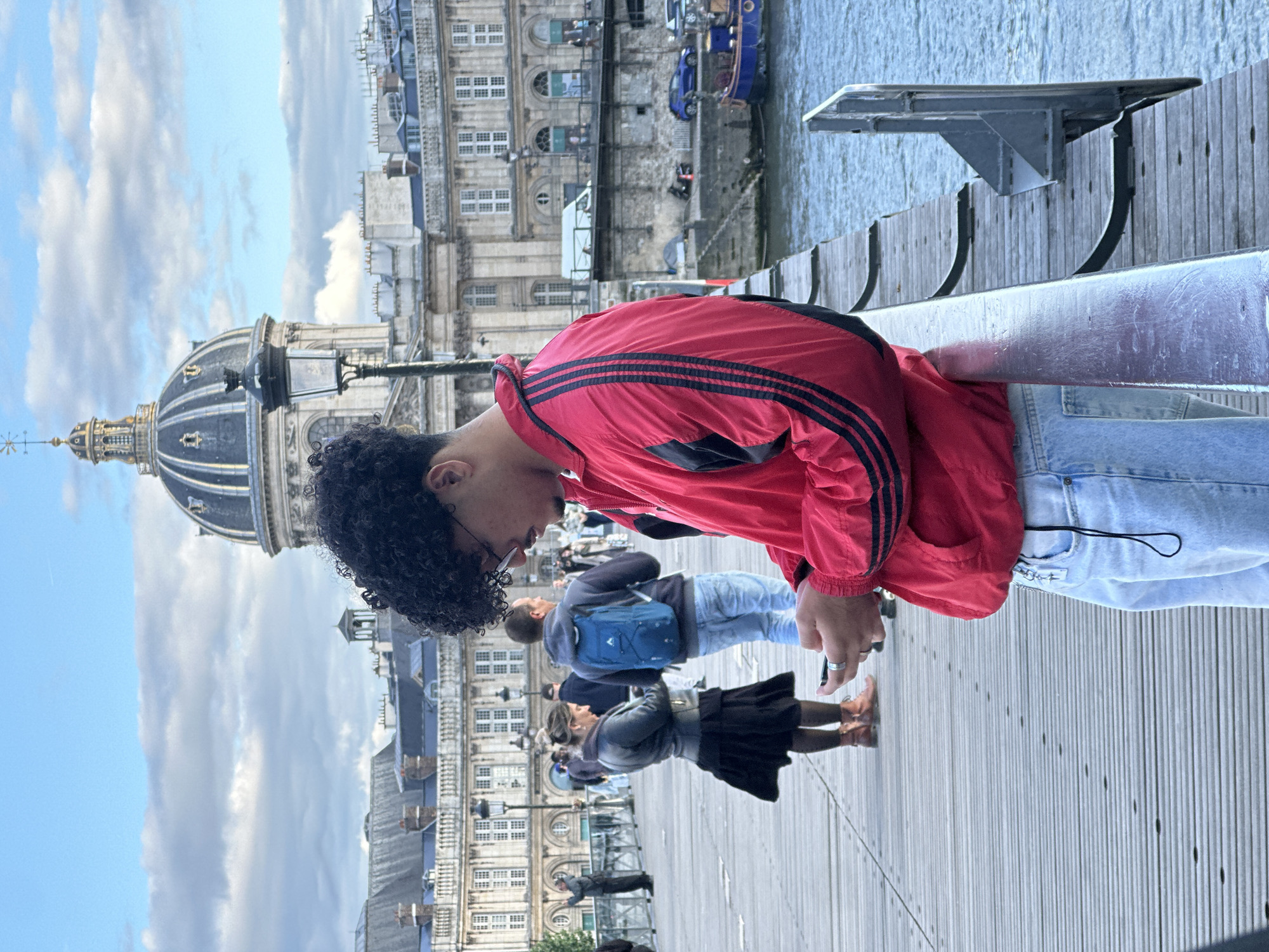 The image captures a man with dark curly hair, wearing a red jacket with black stripes and light blue jeans, standing on a wooden walkway in Paris, France. He is looking down, possibly at a phone or other device in his hands. In the background, there are several other people walking in the same direction. The environment features a large domed building, likely a historic monument, and the Seine River with some boats docked nearby. The weather appears partly cloudy, with a mix of blue sky and clouds. The walkway is lined with a metal railing.