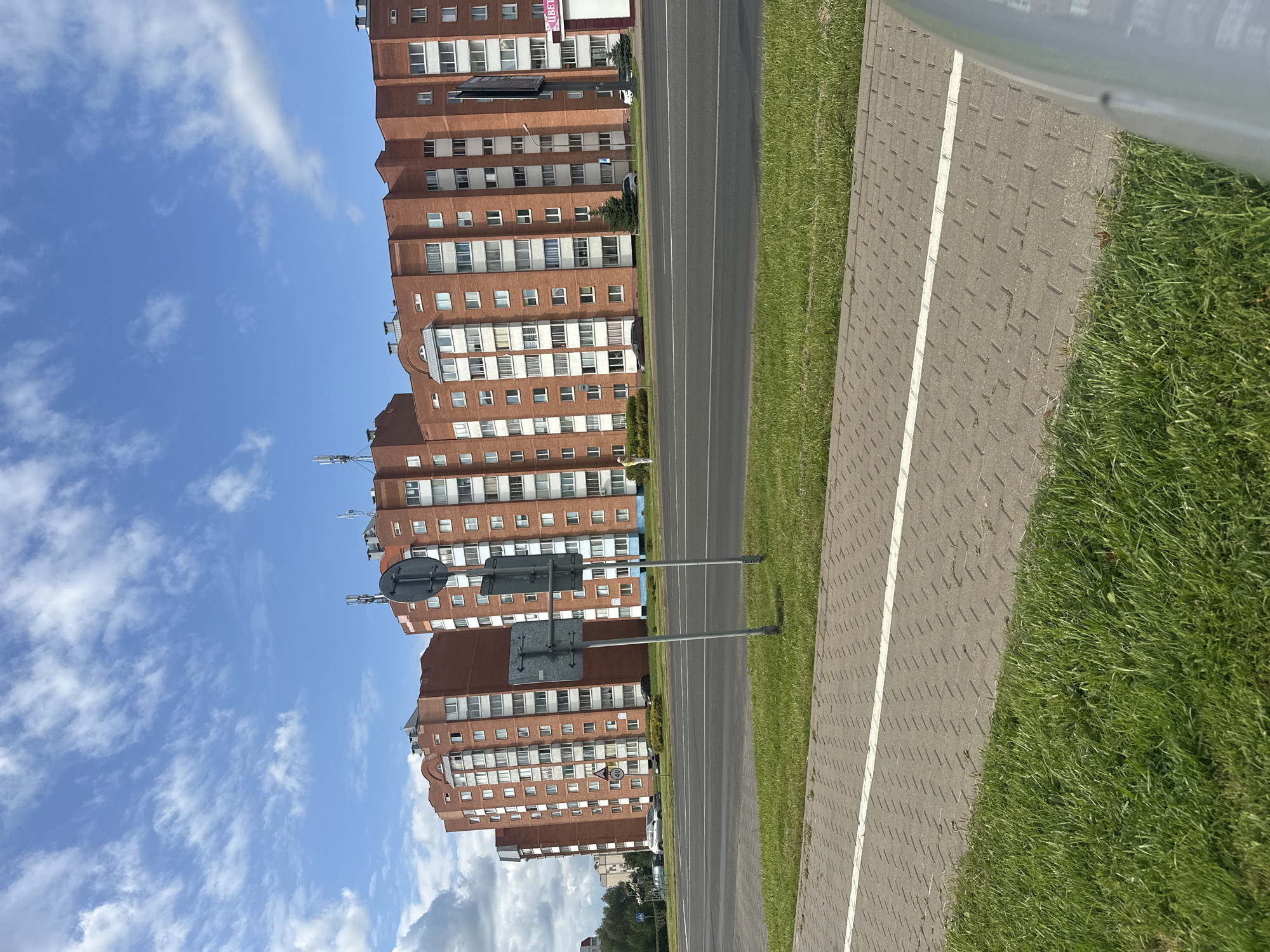 This image, taken in Maladzyechna, Belarus, depicts a wide paved road with a distinct pedestrian walkway adjacent to it. The walkway is constructed of grey interlocking pavers with a white line painted down its center, bordered by a strip of green grass. Across the road, several multi-story brick apartment buildings dominate the skyline. These buildings are characterized by their red brick facades, numerous windows, and visible rooftop antennas. In the background, more greenery and trees are visible, suggesting a semi-urban or suburban environment. The sky above is a bright blue, dotted with white clouds. A few vehicles are visible on the road and parked near the buildings, and street signs are present on the side of the road. The overall scene conveys a typical day in a residential area of the city.