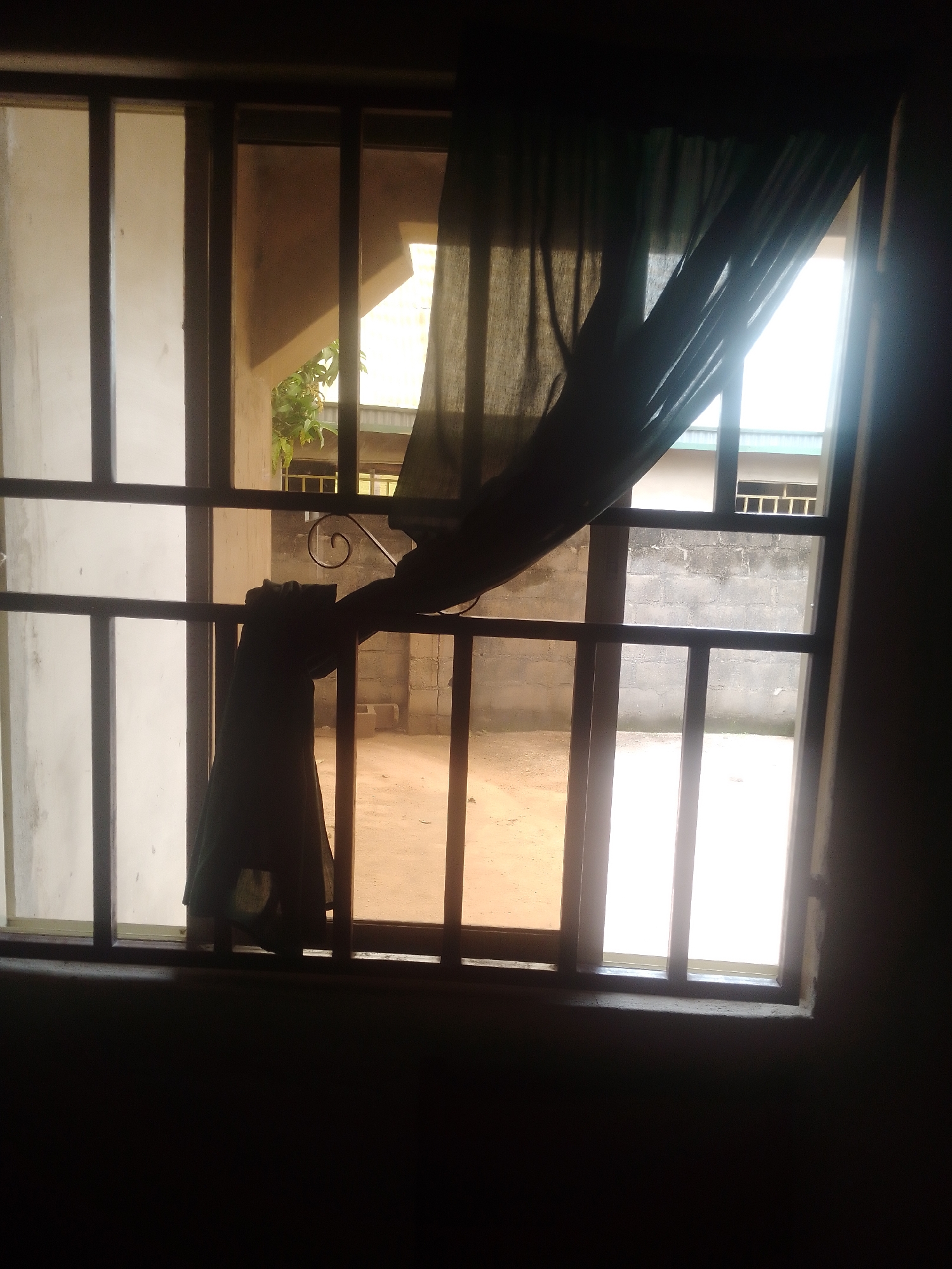 A medium shot captures a view through a barred window looking out into a sunlit courtyard. The window has a dark metal grate with vertical and horizontal bars. A dark green curtain hangs partially drawn, obscuring some of the view and creating shadows. The courtyard outside is paved with dirt or sand, and there is a brick wall in the background. Sunlight streams in from the right, creating bright areas and silhouettes. The overall scene has a somewhat somber or muted tone due to the darkness in the foreground and the limited color palette. The time of day appears to be daytime, likely morning or afternoon, given the strong sunlight. There is no visible text in the image.