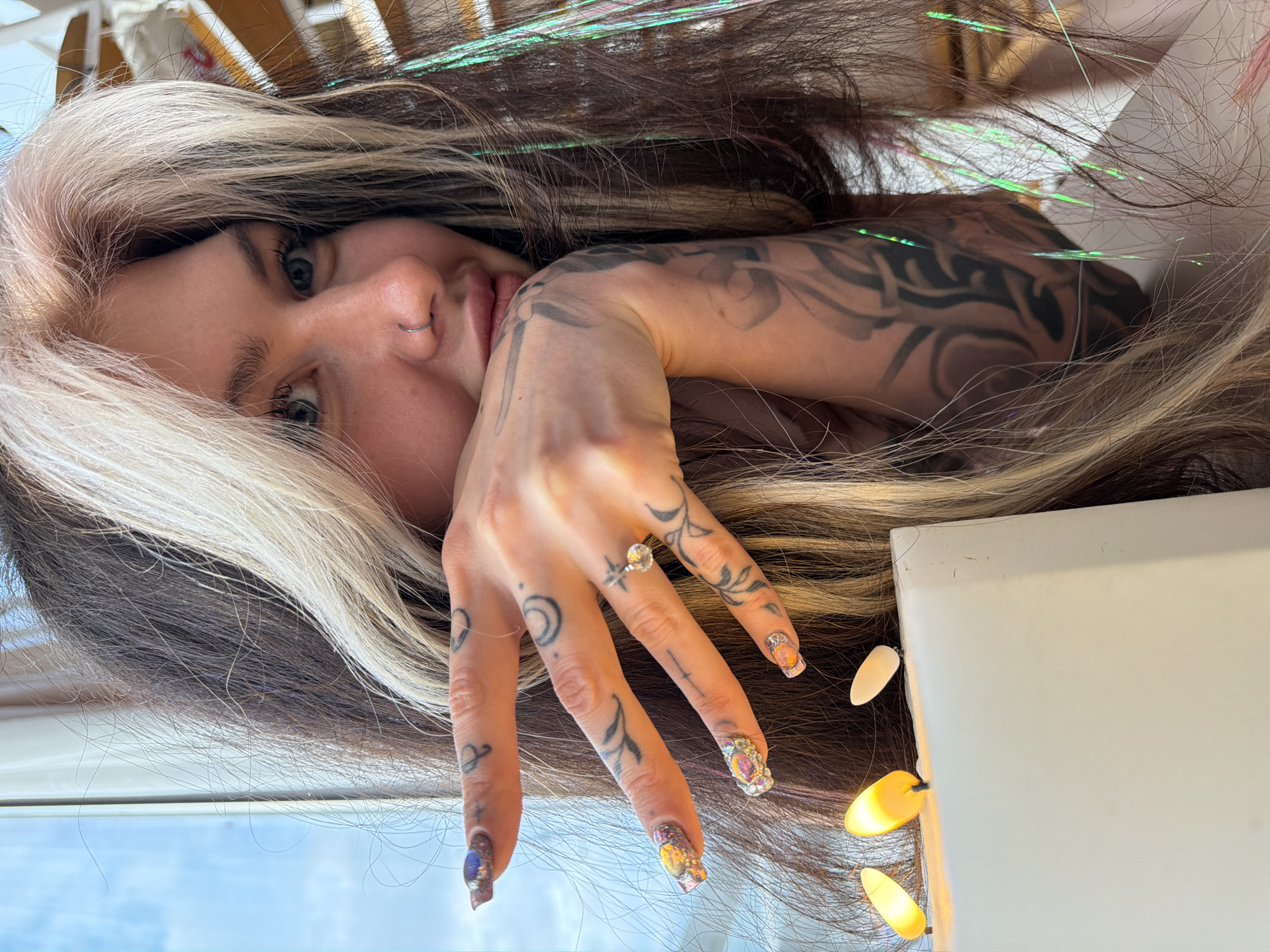 A selfie depicts a woman with dyed brown and blonde hair, a nose ring, and forearm tattoos. She is lying on her back, her left hand outstretched towards the camera, adorned with rings and tattoos. Her nails are decorated with elaborate designs. Beside her hand are a few decorative lights. The background suggests an interior setting, possibly a room in Budva, Montenegro.