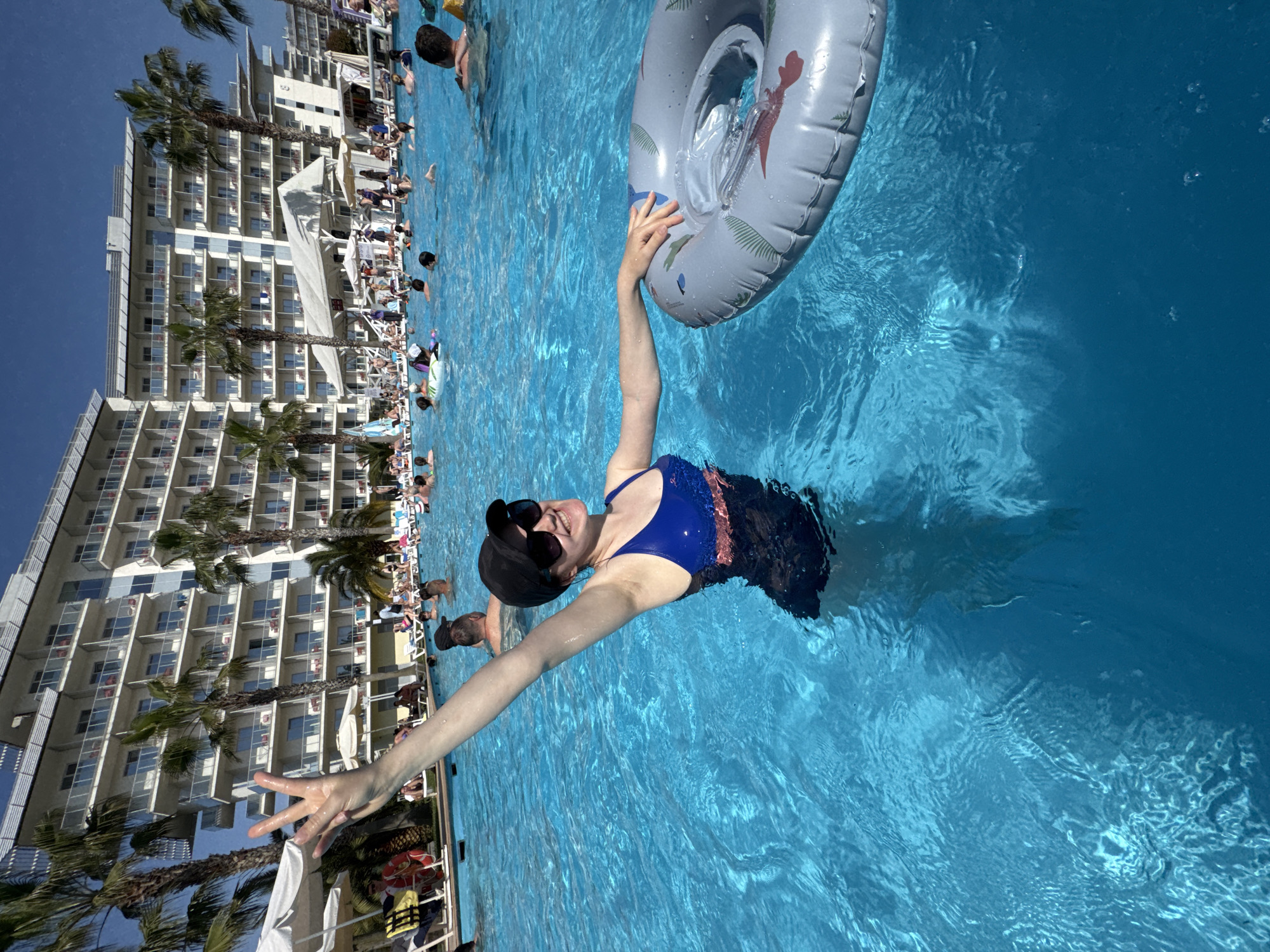 A woman in a blue and black swimsuit is in a swimming pool, holding a gray inflatable tube over her head. She is wearing sunglasses and a black cap. Her left arm is outstretched, fingers splayed. The pool is surrounded by a building with rows of balconies and palm trees, suggesting a resort in a tropical or subtropical climate. Other people are visible in the background, both in the pool and lounging on chairs near the pool's edge. The water in the pool is clear and blue. Sochi, Russia is not confirmed.