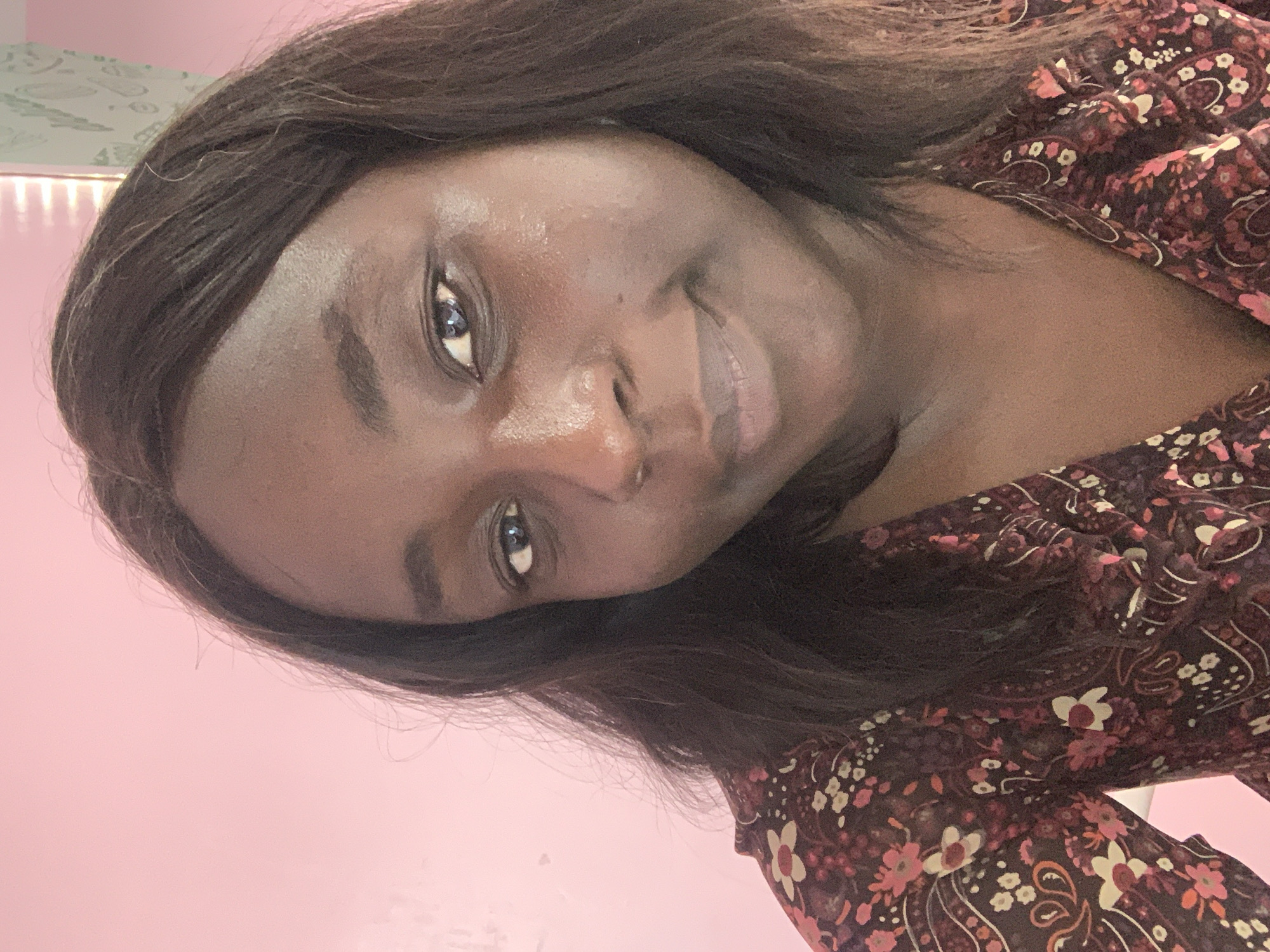 A close-up selfie captures a Black woman in what appears to be an indoor setting. The background is a plain pink wall, with a hint of vertical blinds visible on the right. The woman has dark hair that falls past her shoulders. She is wearing a dark-colored top with a floral pattern featuring pink and white flowers. Her face is angled slightly to the right, and she is looking directly at the camera with a subtle smile. The lighting suggests an overhead light source. The image appears to be taken in Jos, Nigeria.