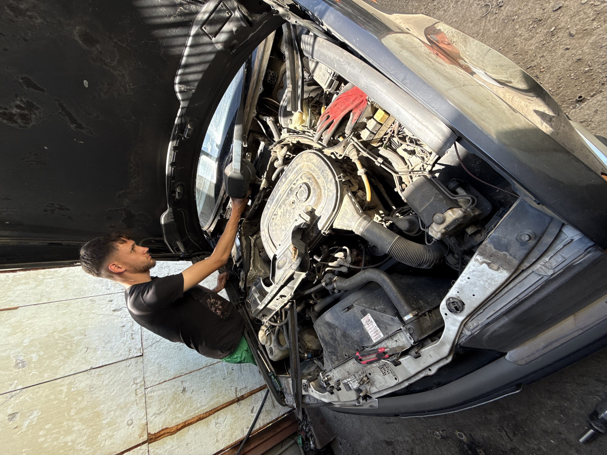 A man is working on the engine of a black car. The hood is open. The engine is exposed and appears to be complex with various hoses and components. The man wears a black t-shirt, green shorts, and appears to be using a tool with a black handle. The scene is indoors, possibly in a garage. The walls are white and textured. The car is parked on concrete. There is no evidence to confirm the location of the city of Shumen, Bulgaria.