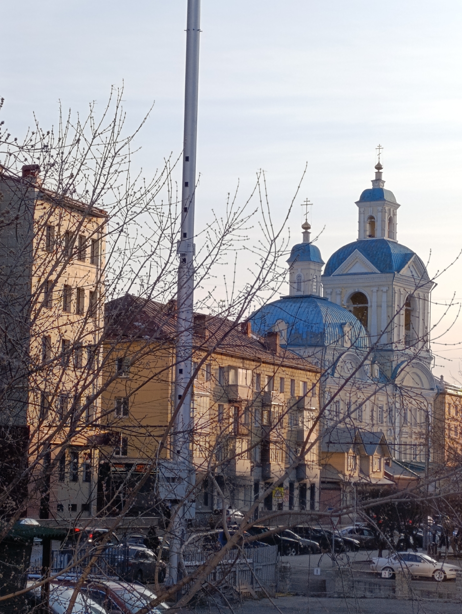 The image shows a cityscape in Krasnoyarsk, Russia, featuring a church with blue domes and a tall bell tower with a cross on top. In the foreground, there are leafless tree branches obscuring the view. Buildings of various heights and colors are visible behind the trees, including a taller building with several windows and a yellow building with a brown roof. A grey pole rises from the lower part of the frame, partially obscuring the buildings behind it. Cars are parked along the street in front of the buildings, and a few people are discernible in the distance. The sky is overcast, giving the scene a muted tone.