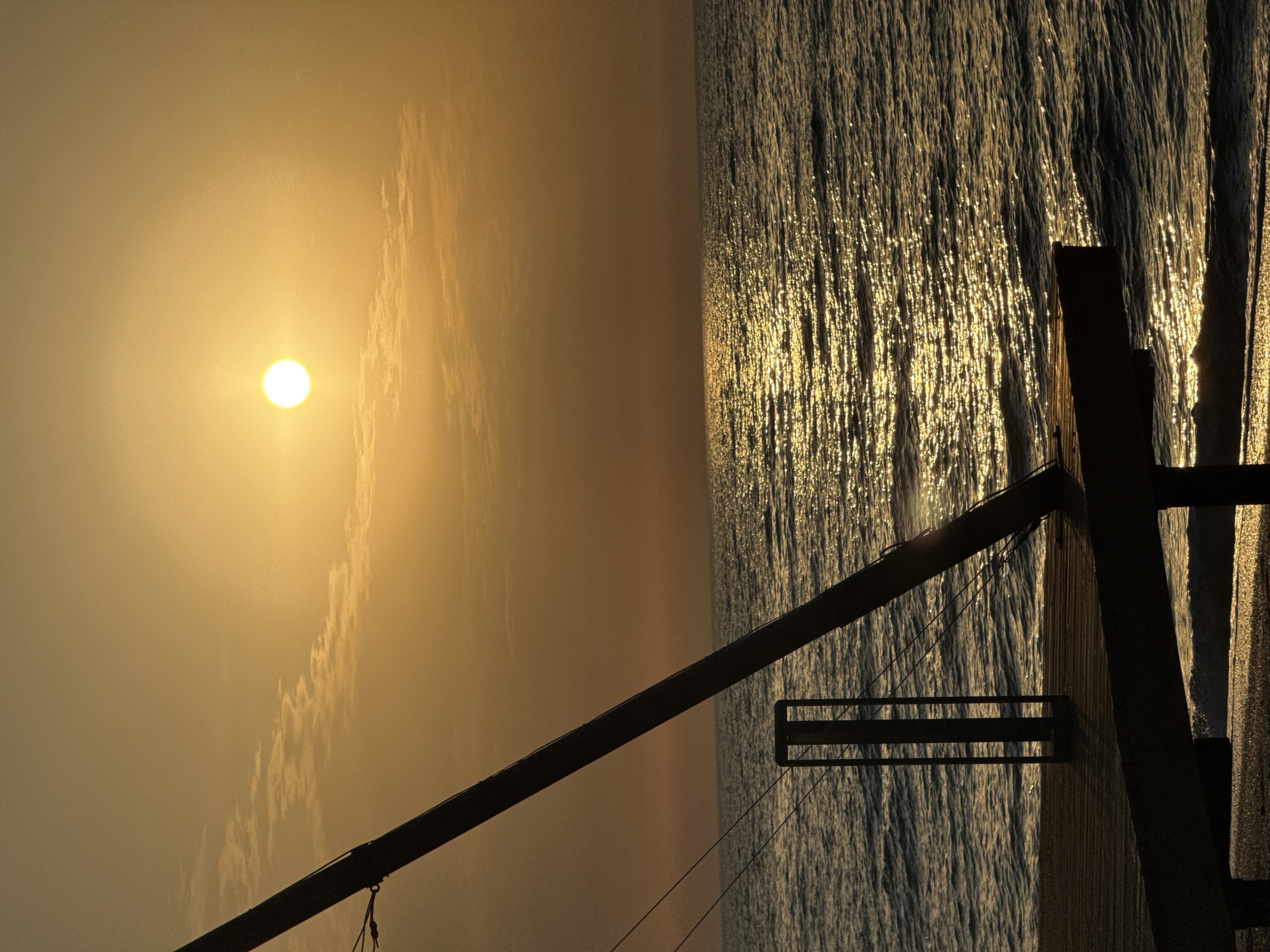 The image depicts a sunrise or sunset scene, likely near the coast of Мысовское сельское поселение, Ukraine. The sun is visible in the sky, casting a warm, golden glow. The sea reflects the light, creating a shimmering effect. A wooden structure, possibly a pier or dock, is silhouetted against the water. The sky is hazy, and some clouds are present.