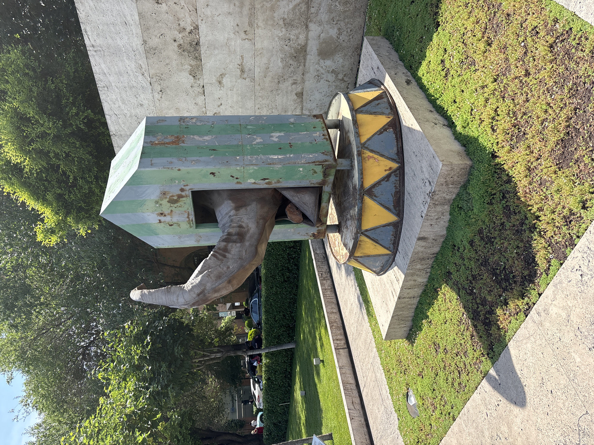 An outdoor sculpture of an elephant's head and trunk emerges from a green and grey striped, weathered metal structure resembling a small building. The structure sits on a circular base decorated with yellow and blue triangles, which is itself placed on a concrete pedestal. The sculpture is situated in a green lawn area with paved pathways and surrounded by trees. In the background, there is a large concrete wall and some parked cars and people on a street. This sculpture is located in Yerevan, Armenia, specifically at the Cascade complex.