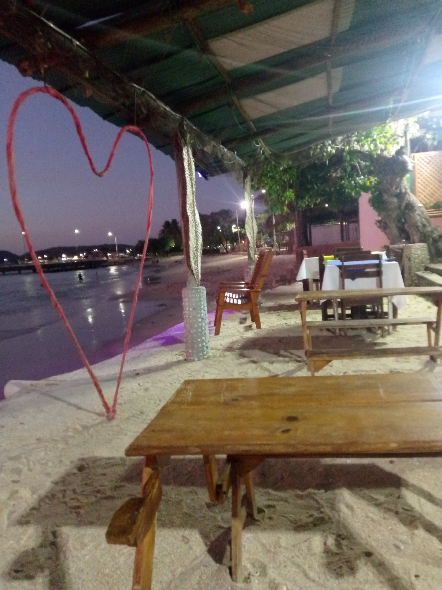 The scene captures an open-air restaurant at dusk, likely located on the beach in Juan Griego, Venezuela, suggested by the nearby ocean and lights across the water. A large, roughly-hewn wooden table occupies the foreground, its surface showing wear. Behind this table are other wooden tables and chairs, some with white tablecloths, sparsely populated. A prominent red heart-shaped decoration hangs from the structure above. The beach and ocean are visible in the background, with faint lights indicating activity or buildings across the bay. The overall atmosphere is relaxed, possibly indicating a quiet evening.