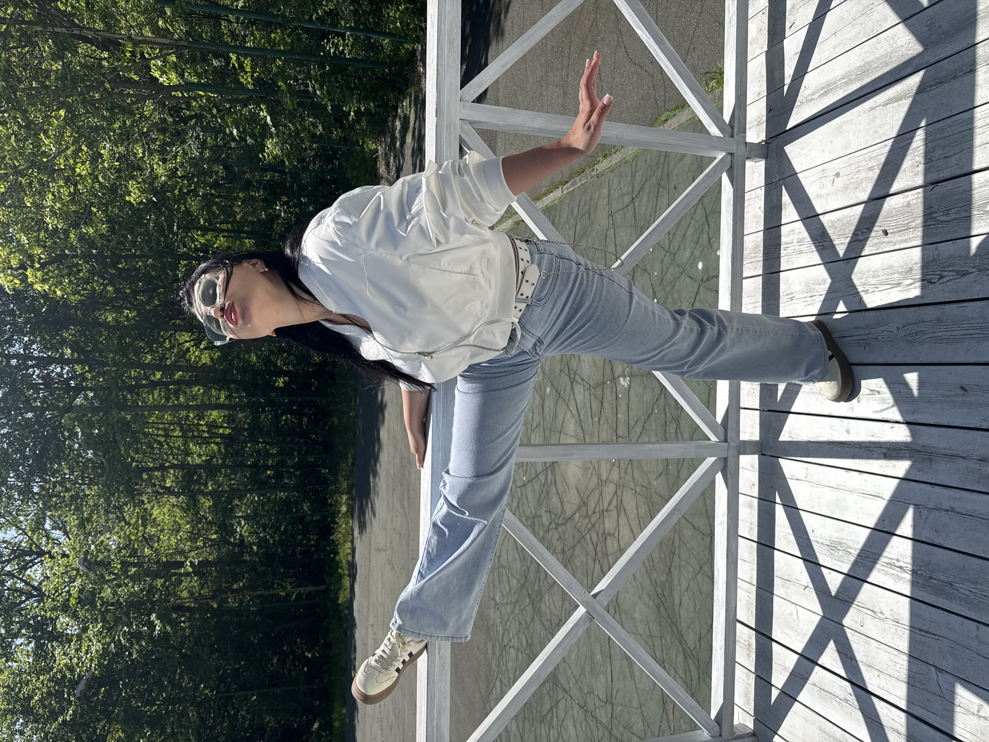The photograph depicts a woman with dark hair wearing sunglasses, a white jacket, and light blue jeans. She is standing on a white wooden structure that appears to be part of a bridge or raised platform. The structure casts geometric shadows on the wooden planks below. The background shows green trees suggesting a park or forested area. The scene is brightly lit, with the sun creating stark contrasts in light and shadow. While it's not possible to confirm the location, the description would remain unchanged regardless.
